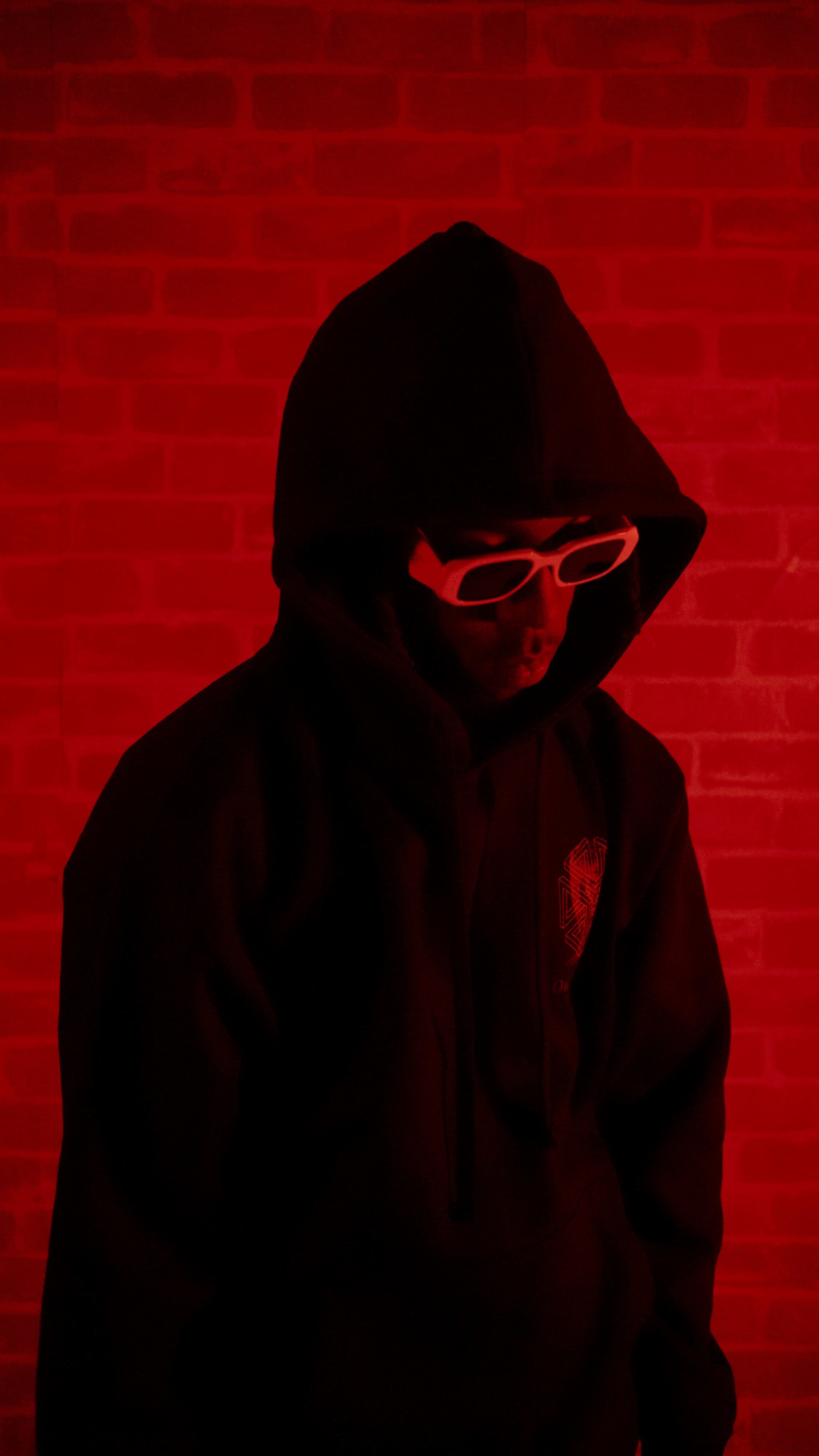A person wearing a black hoodie and white sunglasses stands against a red brick wall.