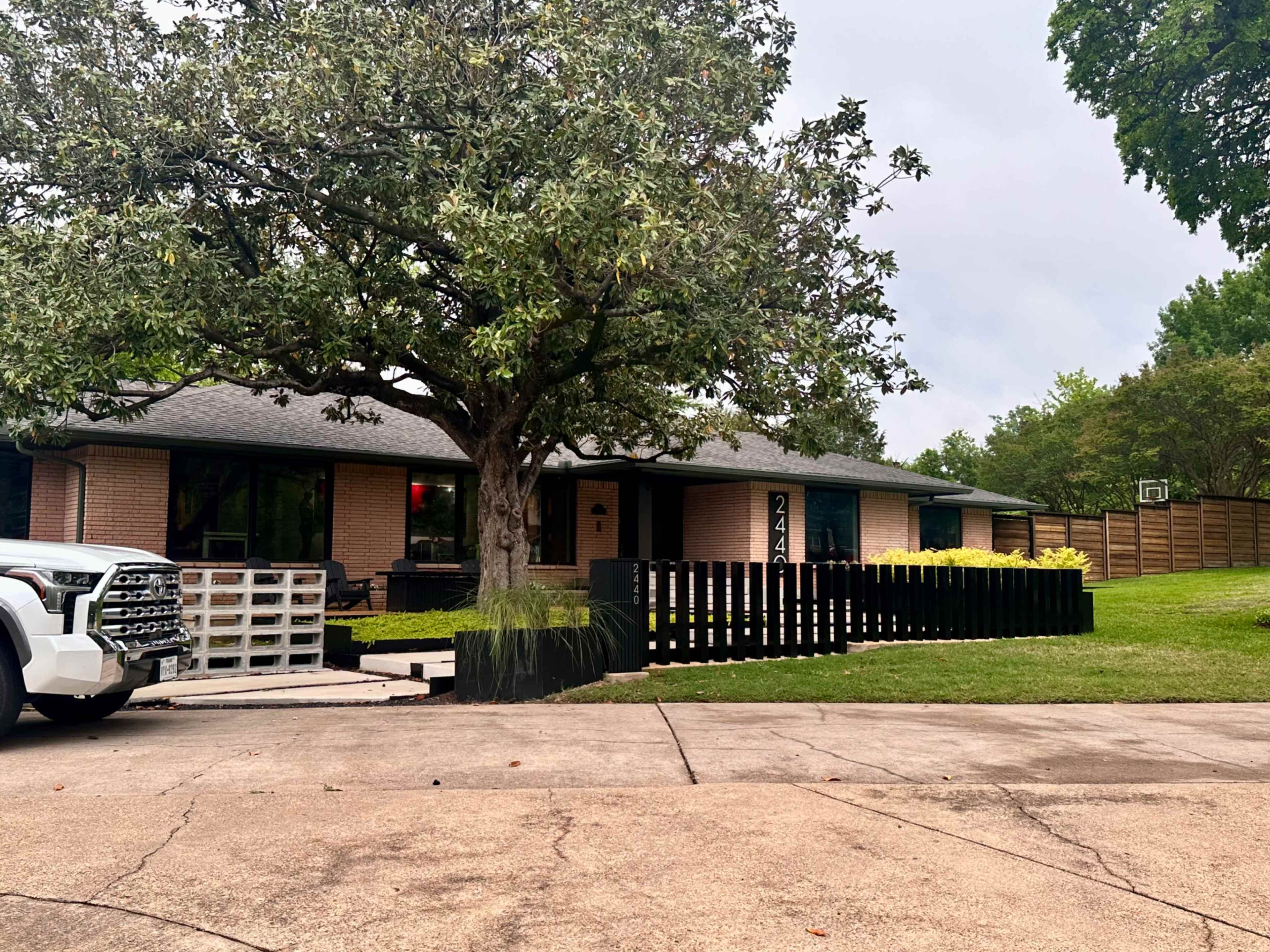 The image shows a single-story house with a large tree in the front yard and a black wooden fence enclosing the property.
