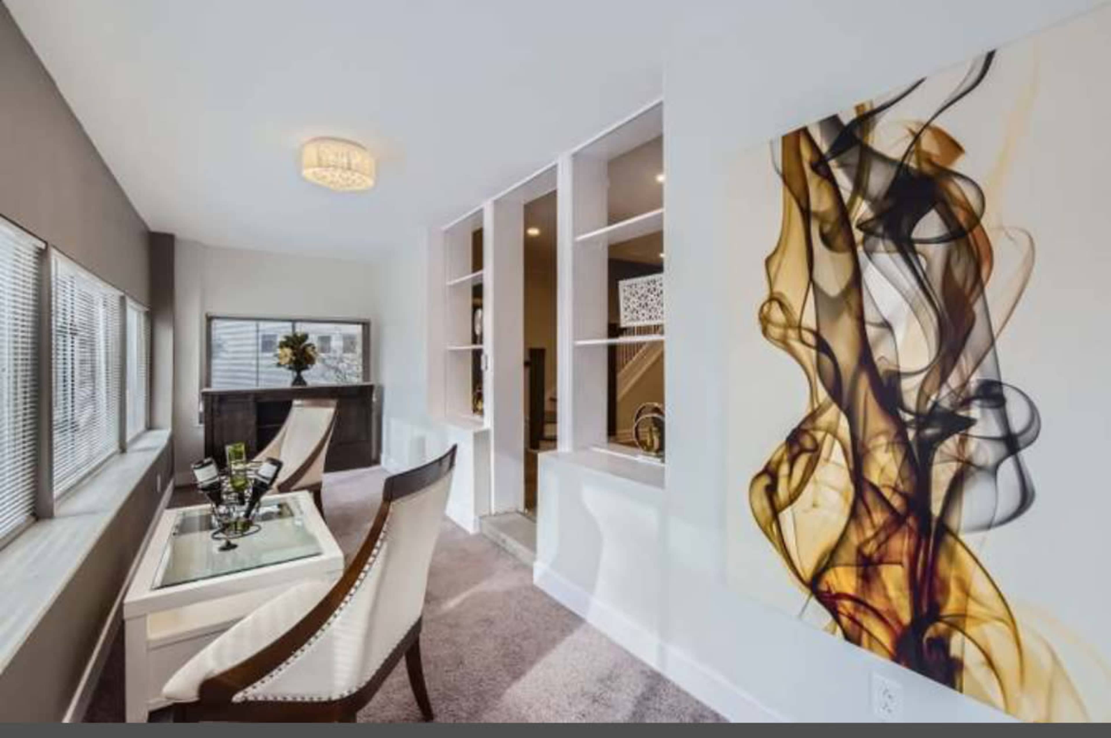 The image shows a modern interior space featuring a glass table with two chairs, an abstract wall art piece, and shelves, all illuminated by natural light through large windows.