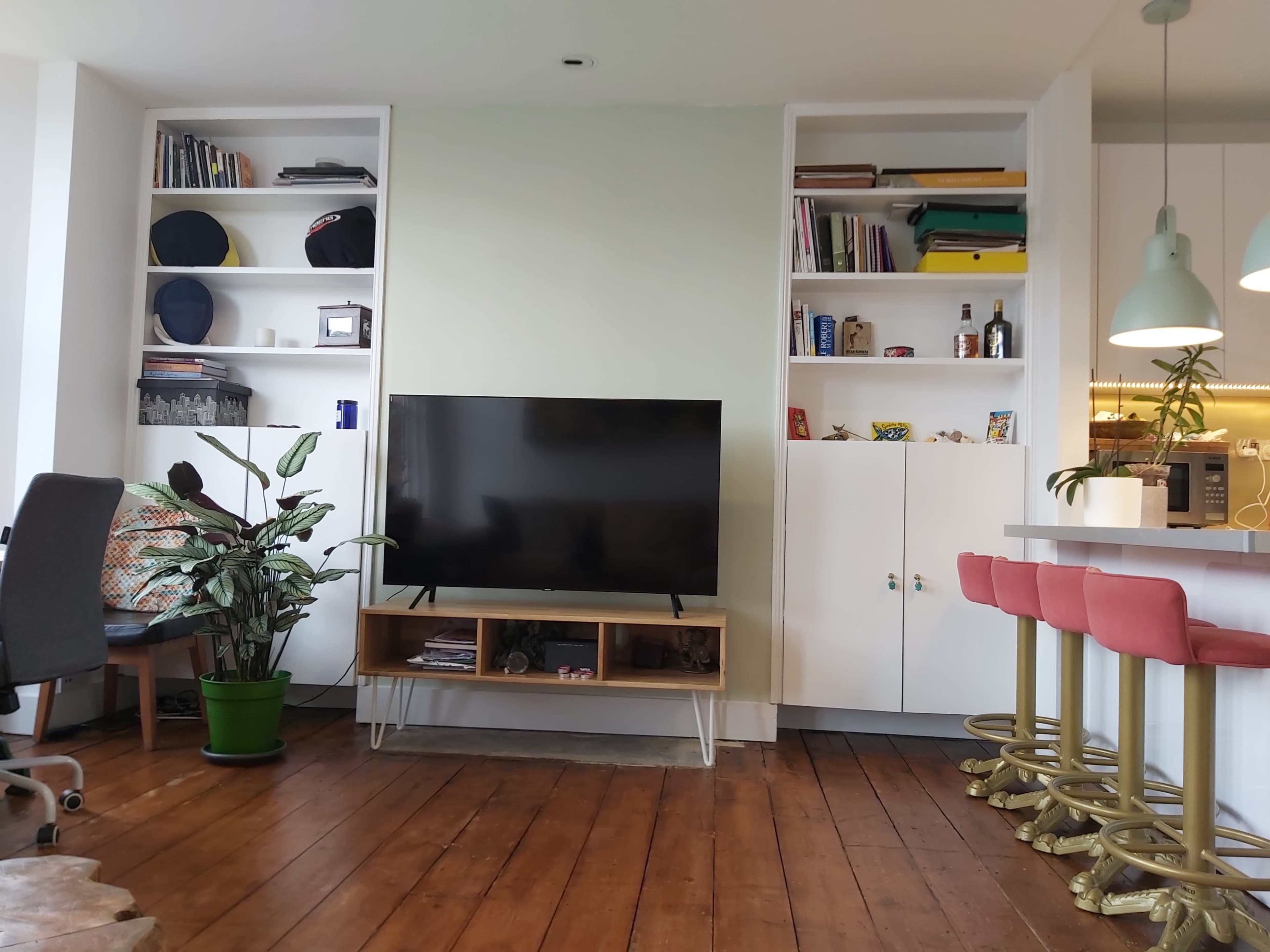 A living room features a television on a wooden stand, surrounded by shelves with books and decorative items, alongside a green plant in a pot and a bar with pink stools.