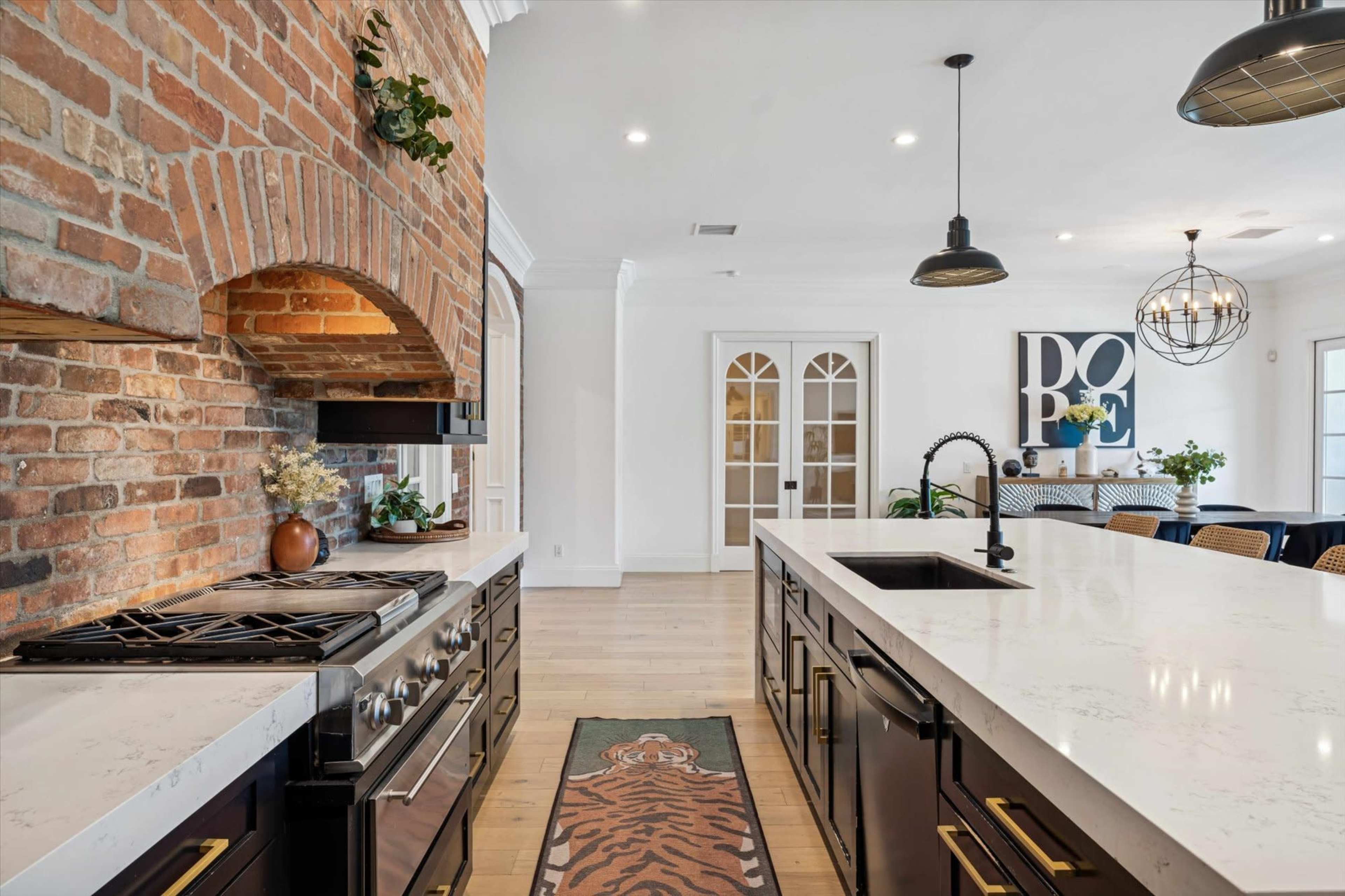 A modern kitchen features a brick arch over a gas stove, with dark cabinetry, a white countertop, and a dining area visible in the background.