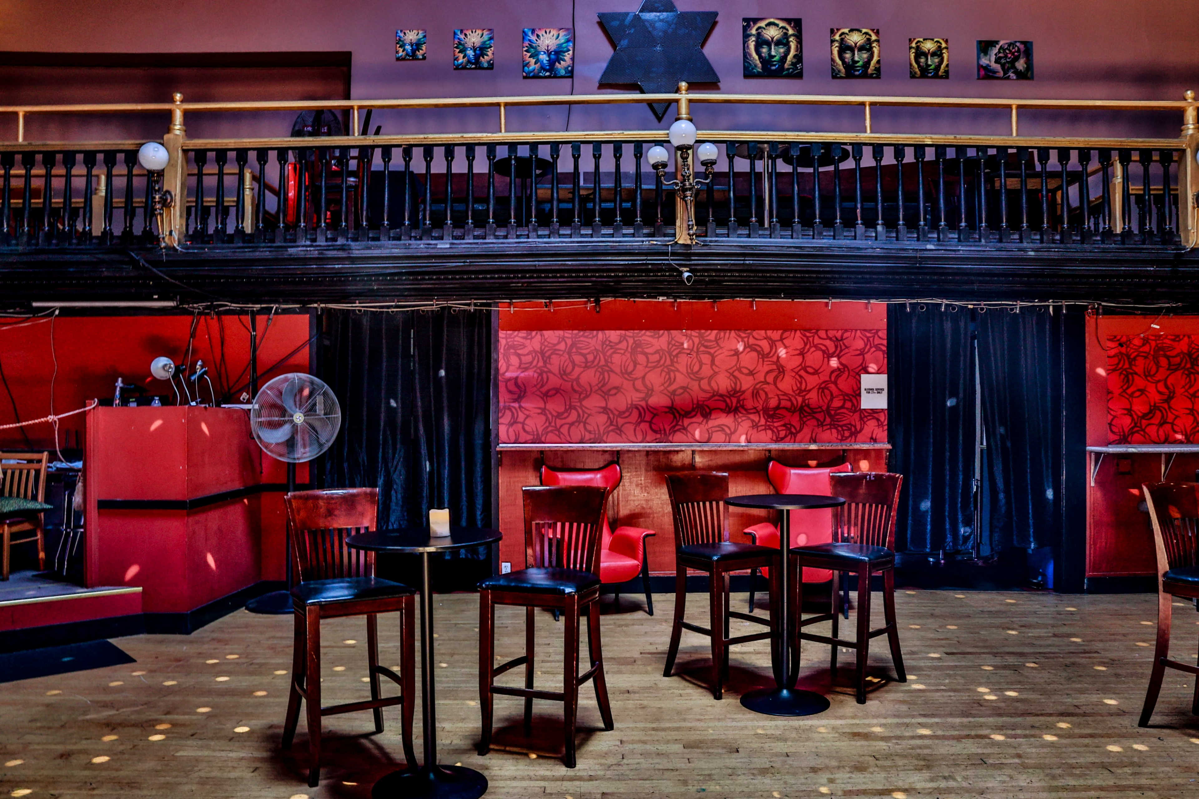 The interior of a dimly lit bar features empty wooden tables and chairs on a wooden floor, with red walls adorned by artwork and a balcony above.
