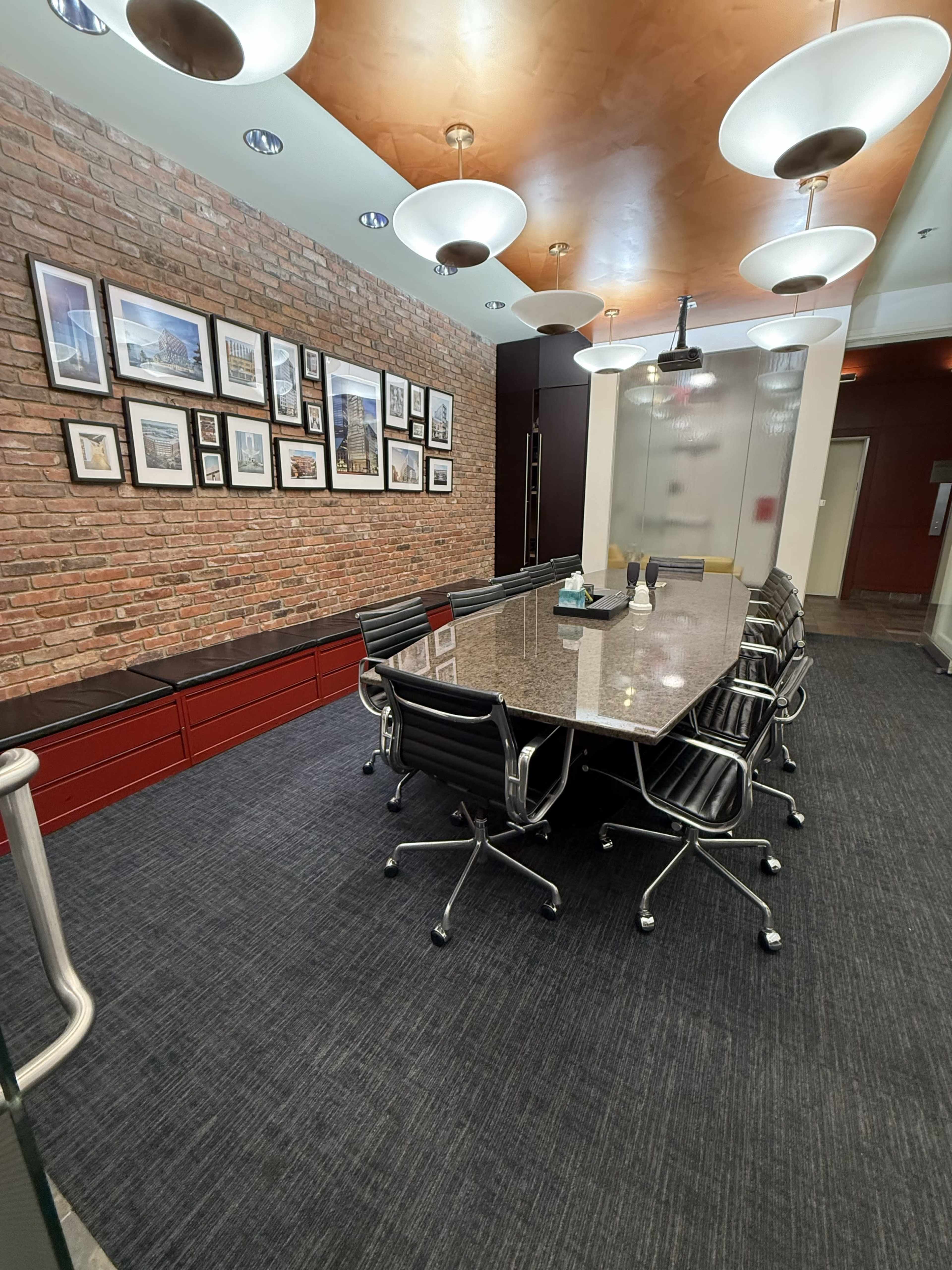 A modern conference room features a large rectangular table surrounded by black ergonomic chairs, with a brick wall displaying framed artwork and two overhead light fixtures.