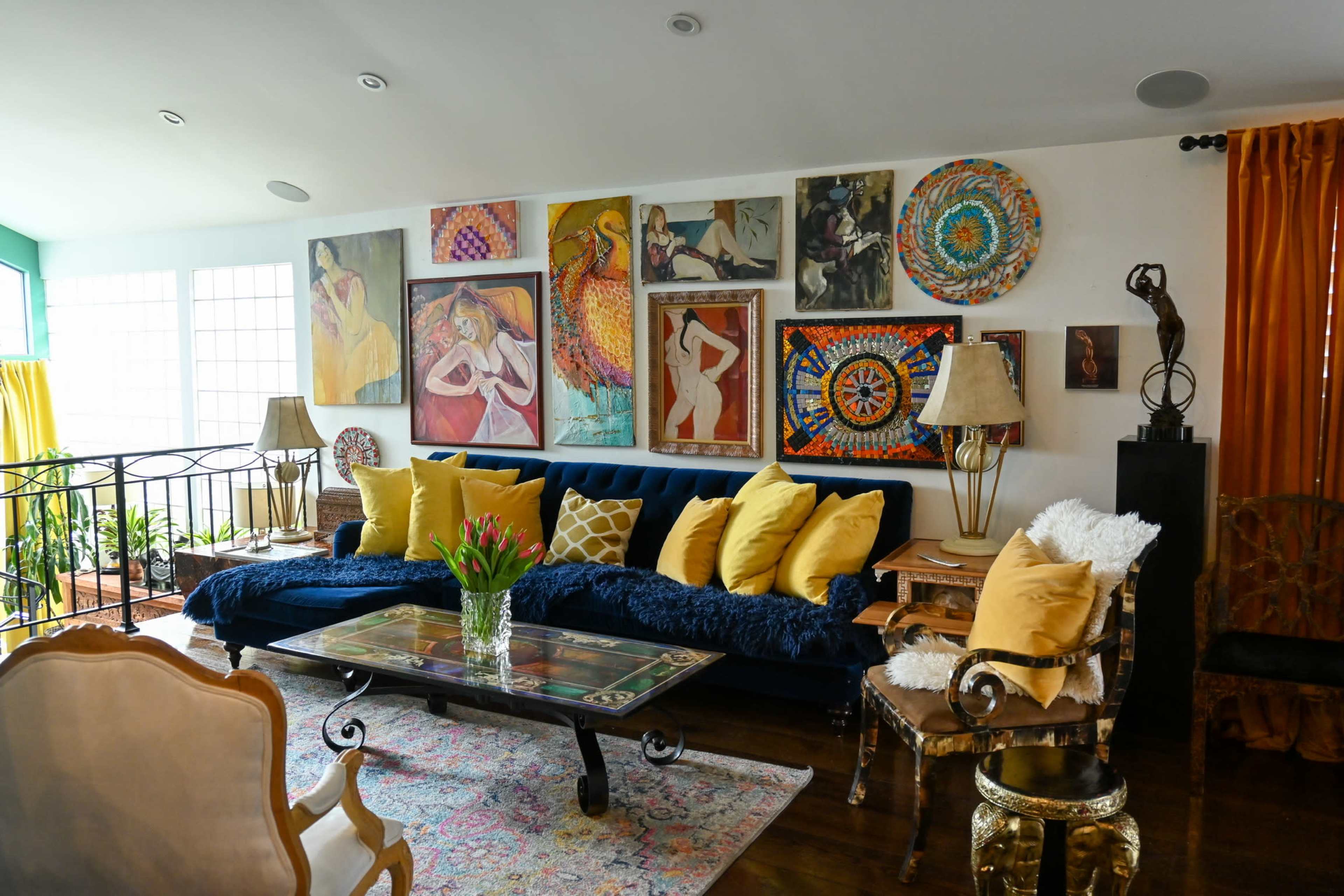 A cozy living room features a blue sofa adorned with yellow pillows, a glass-topped coffee table, and various artworks hanging on the walls, along with a mix of decorative furniture.