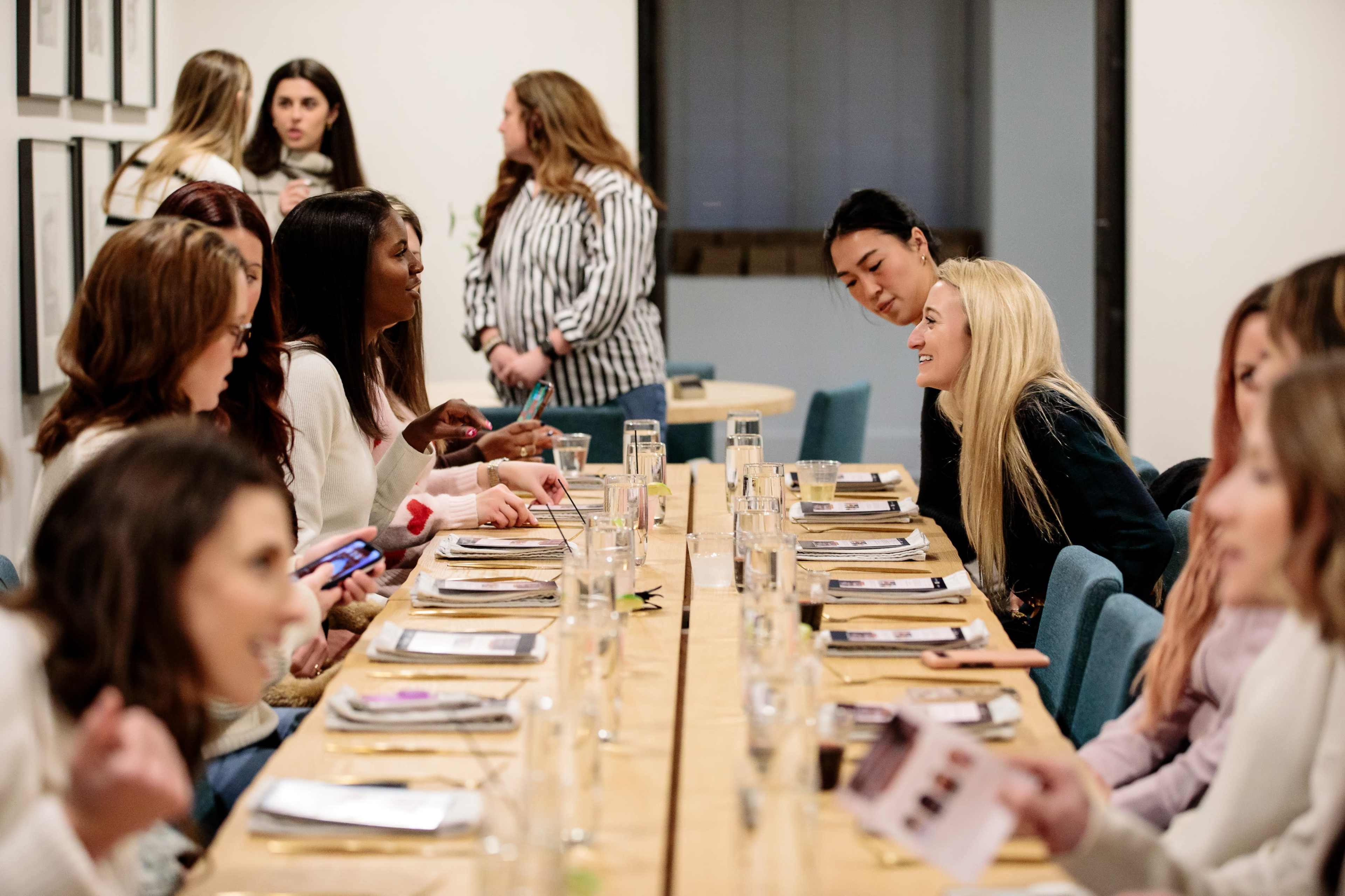 A group of women is gathered around a long wooden table in a well-lit room, engaged in conversation while plates and glasses are set for a meal.