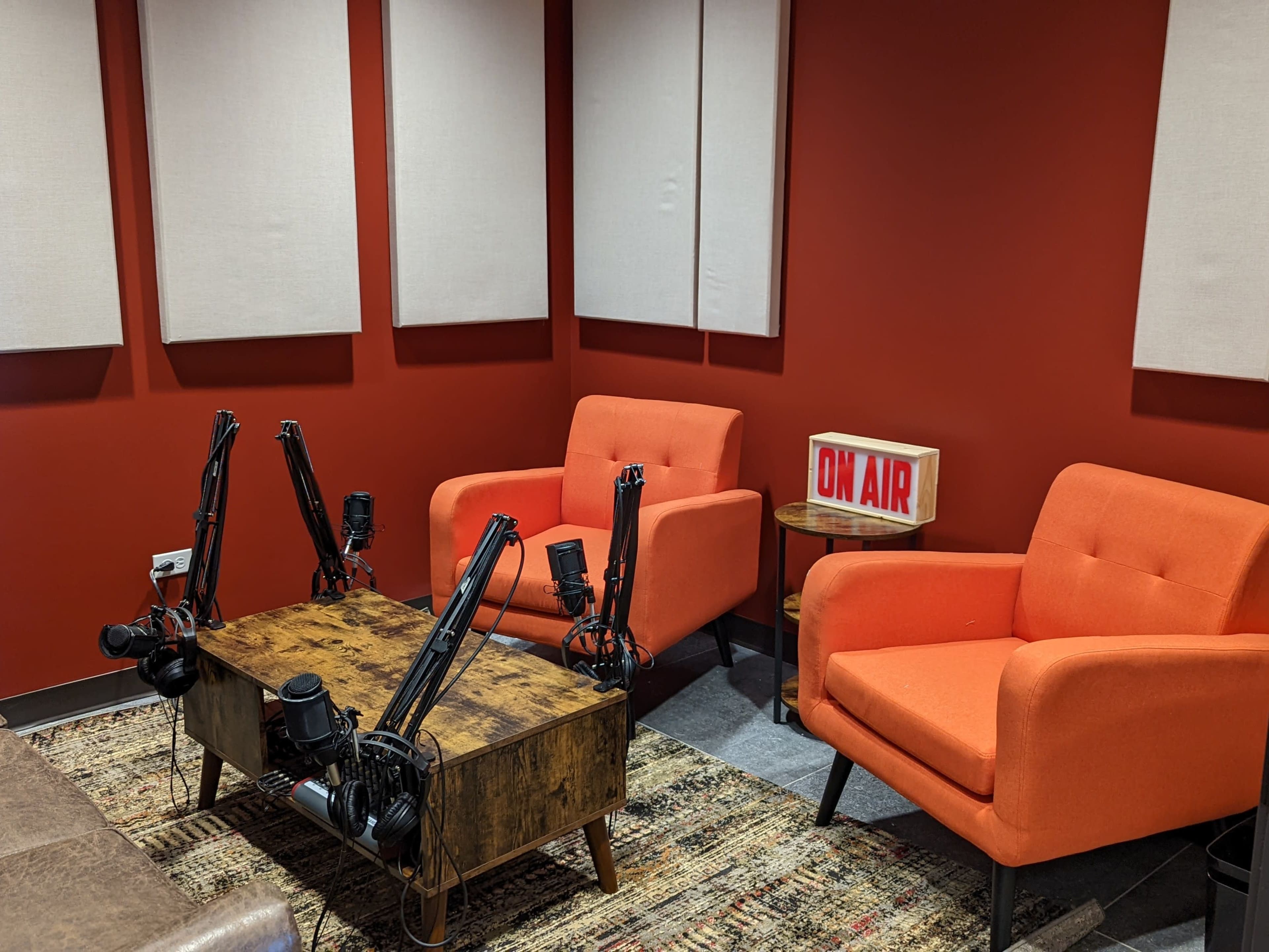 A recording studio with two orange chairs, a wooden table, and a lit "ON AIR" sign against a red wall.