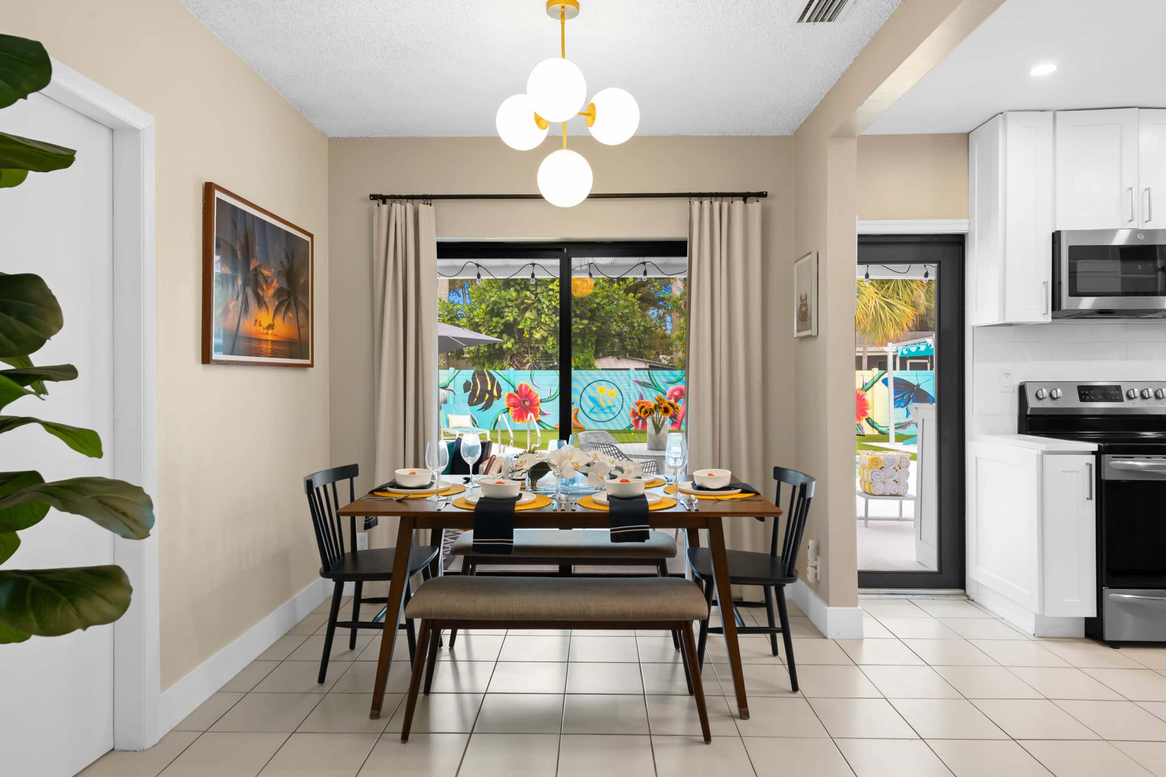 A dining area with a table set for a meal is visible, featuring black chairs, a bench, and large windows overlooking a colorful outdoor space.
