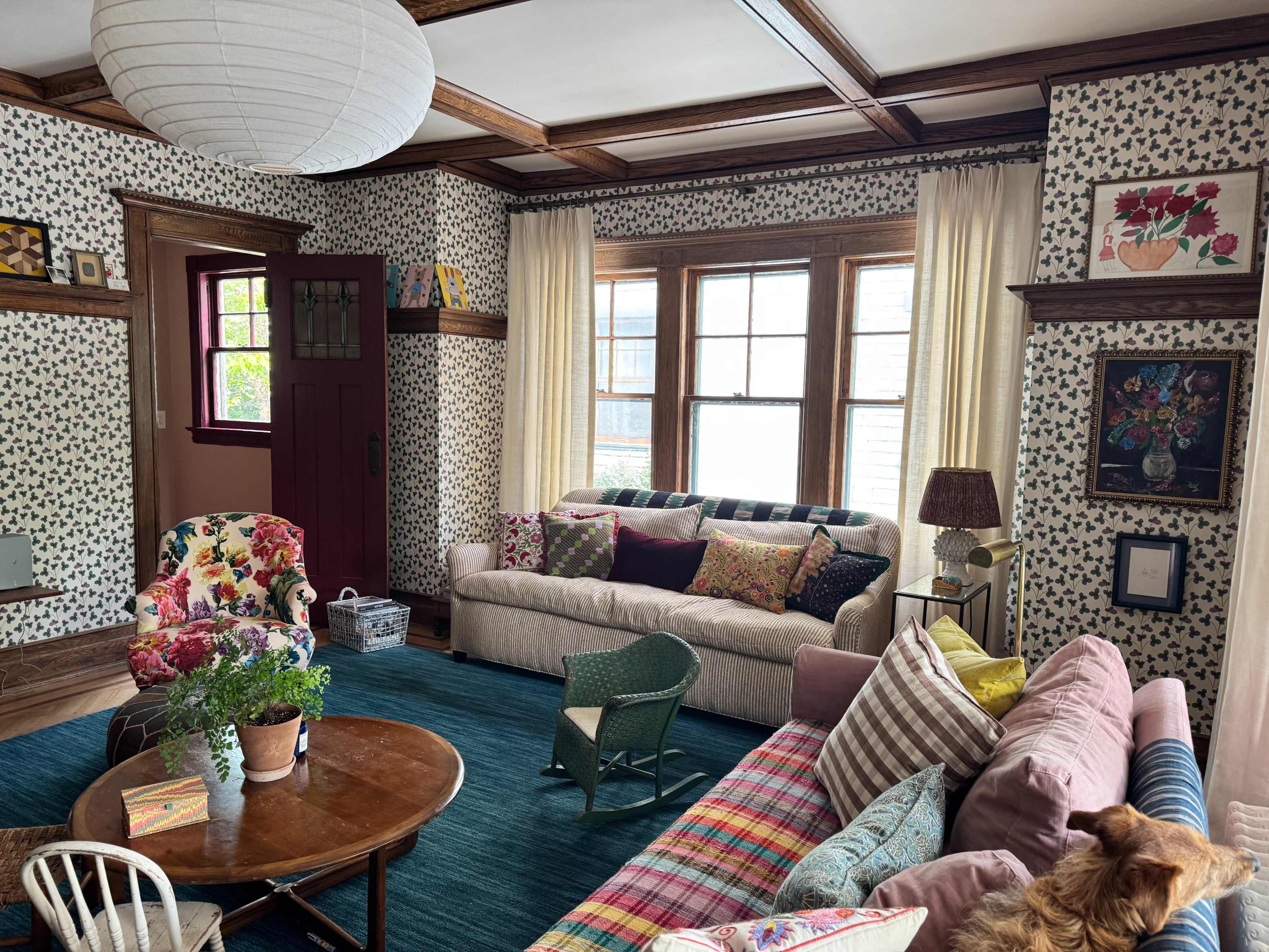 The living room features a mix of colorful furniture and patterned wallpaper, with a round wooden table at the center and large windows providing natural light.