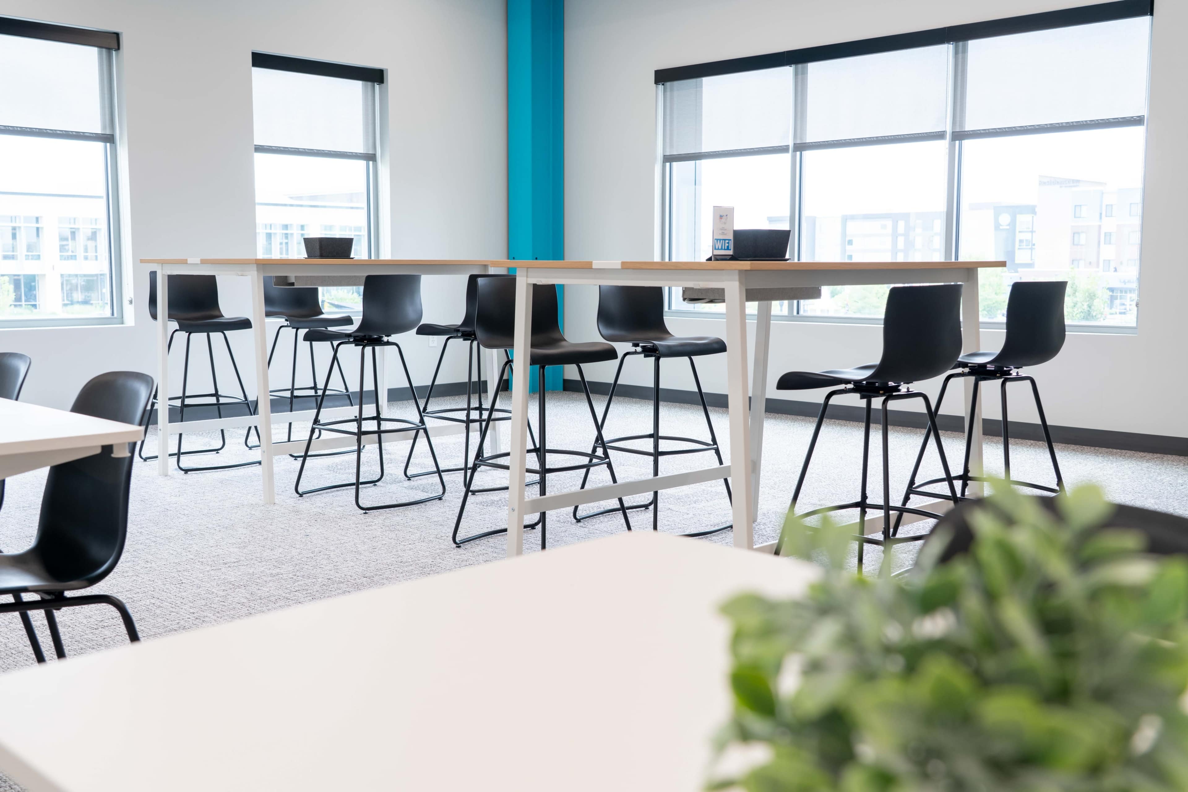 The image shows a modern workspace with tall tables and black chairs arranged near large windows.