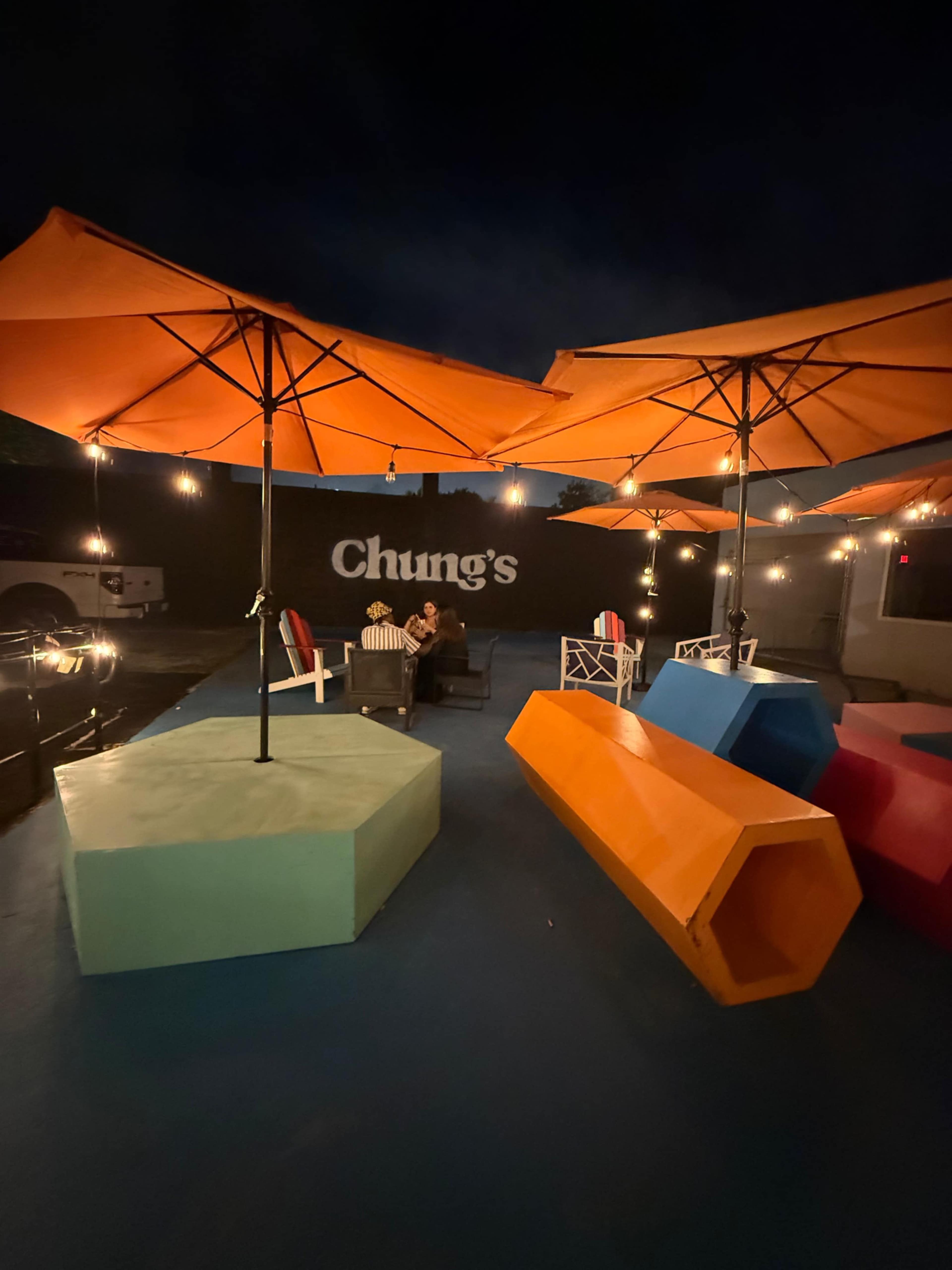 The image shows a dimly lit outdoor area with colorful geometric seating, large orange umbrellas, and a wall displaying the name "Chung's."
