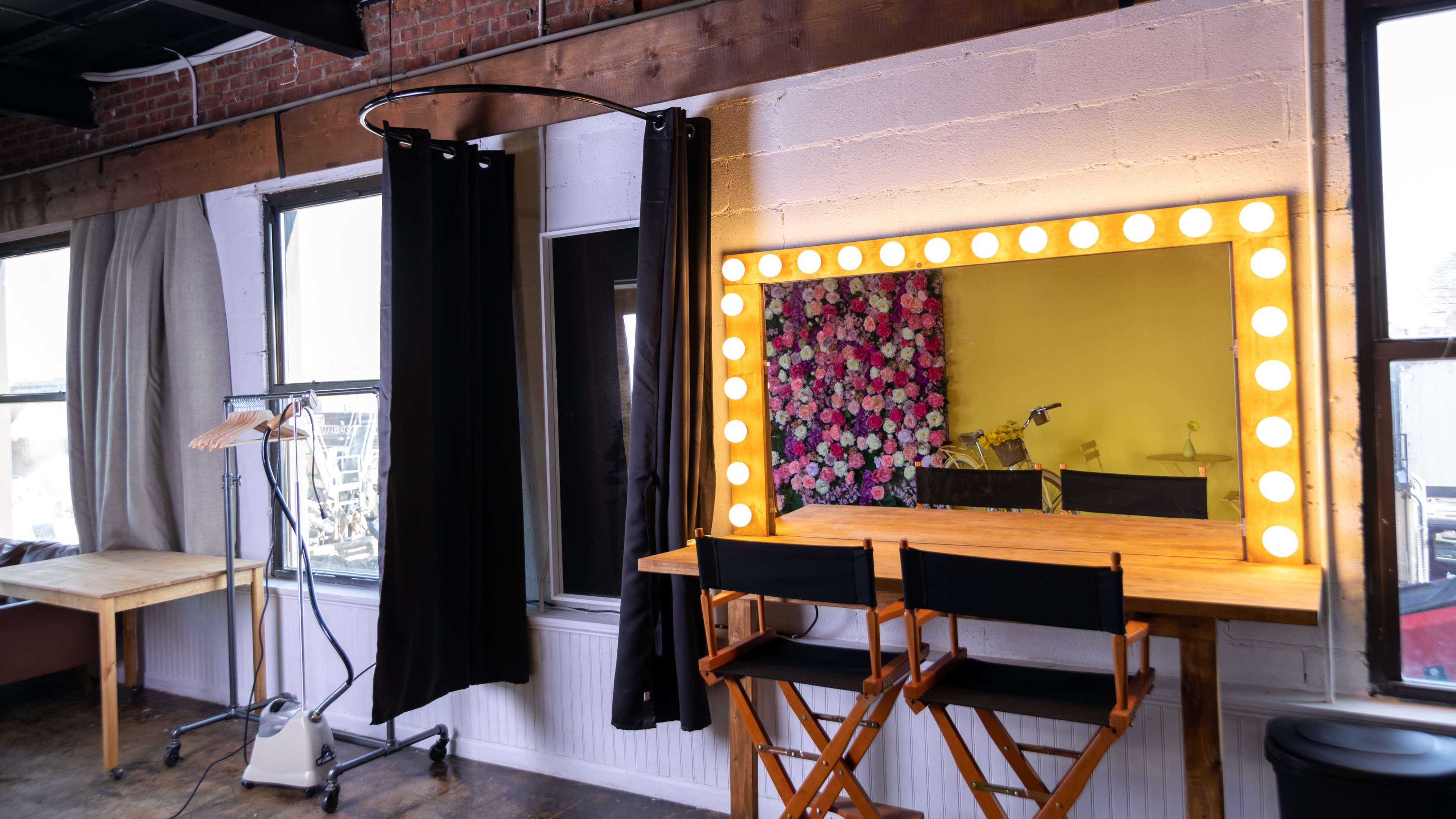 The image shows a bright room with a large mirror framed by light bulbs, a clothing rack, and two director's chairs in front of the mirror.