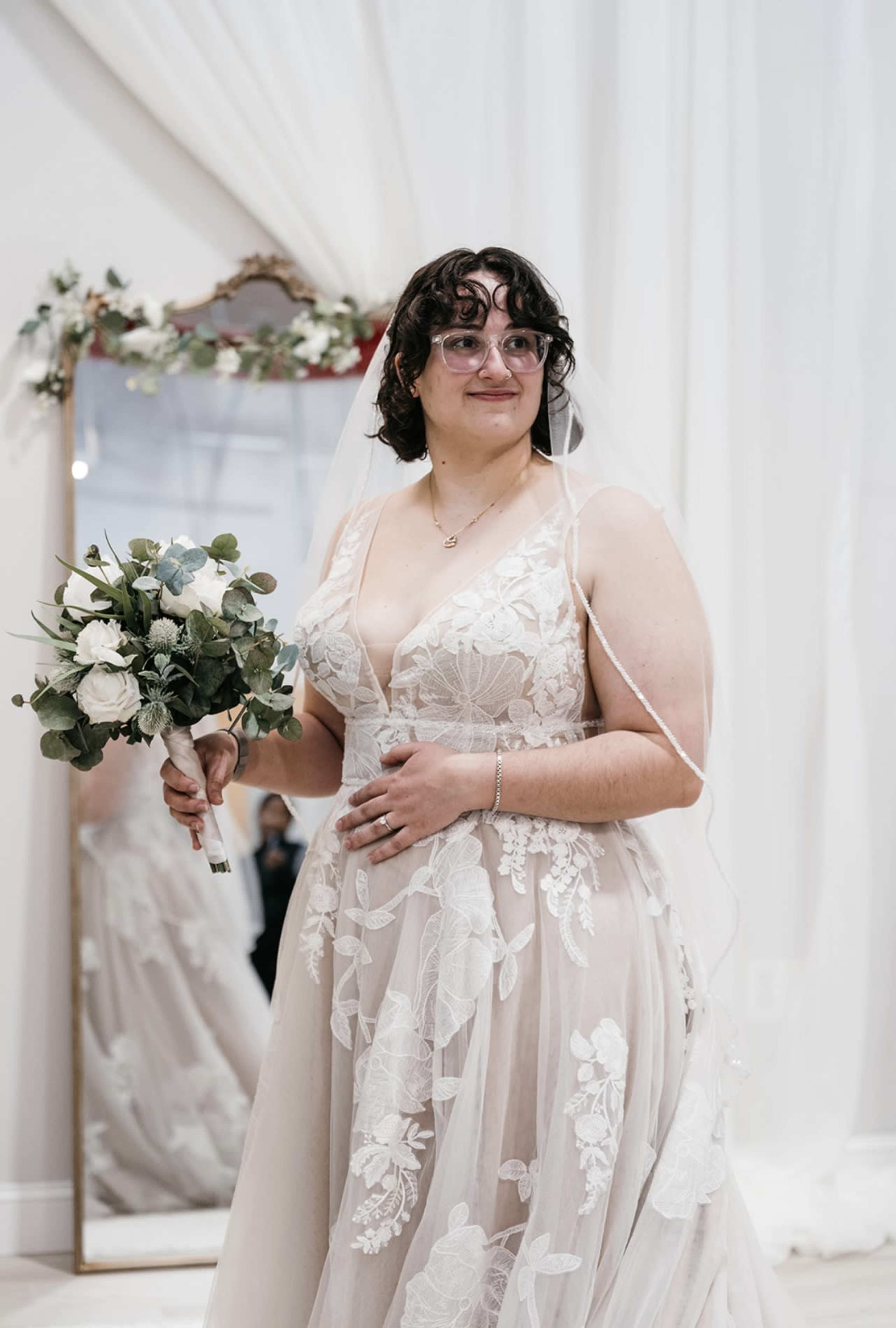 A person in a wedding dress holds a bouquet of flowers and stands in front of a mirror adorned with floral decorations.
