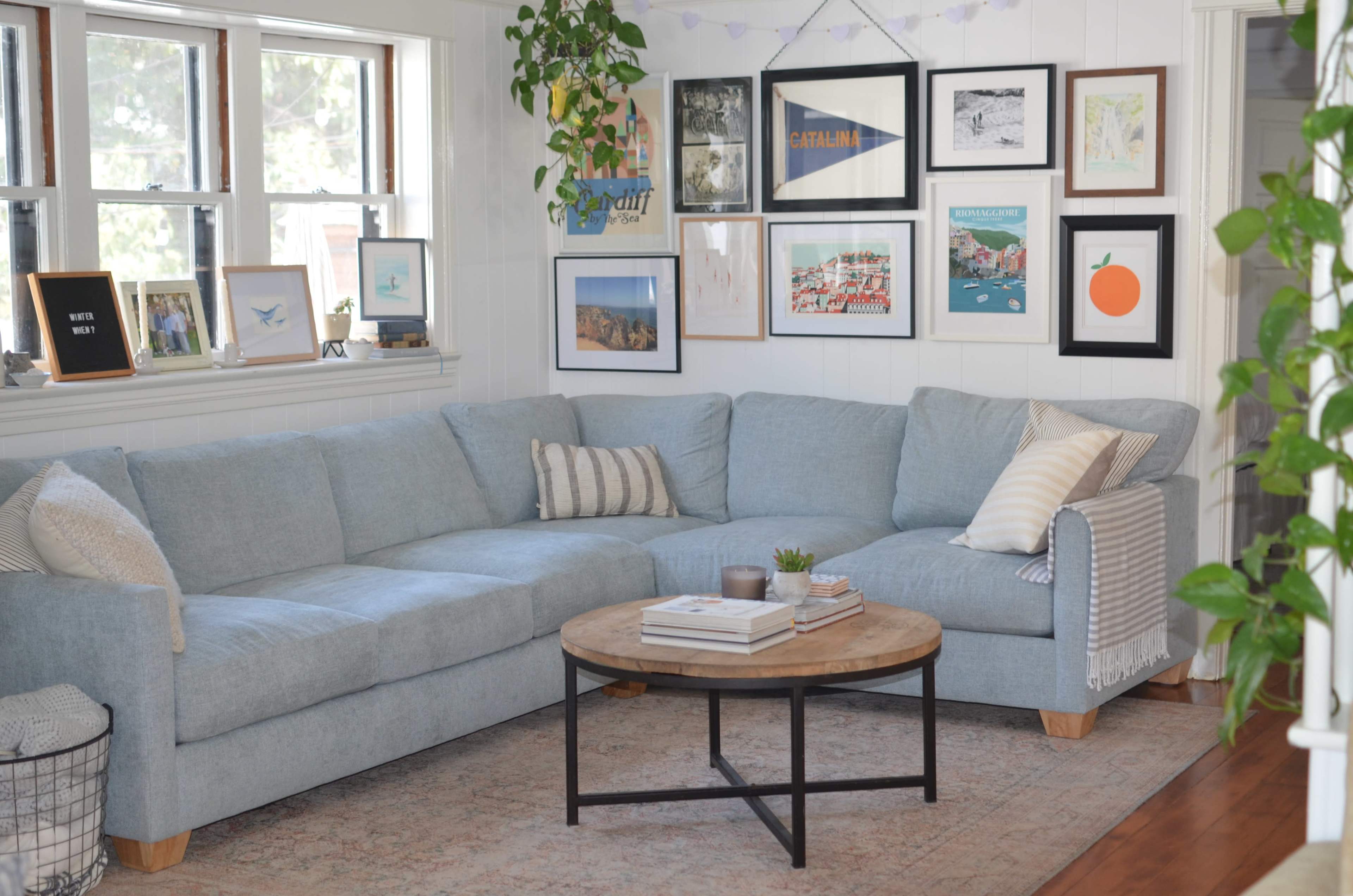 The image shows a cozy living room with a large light blue sectional sofa, a circular wooden coffee table, and various framed artworks on the walls.