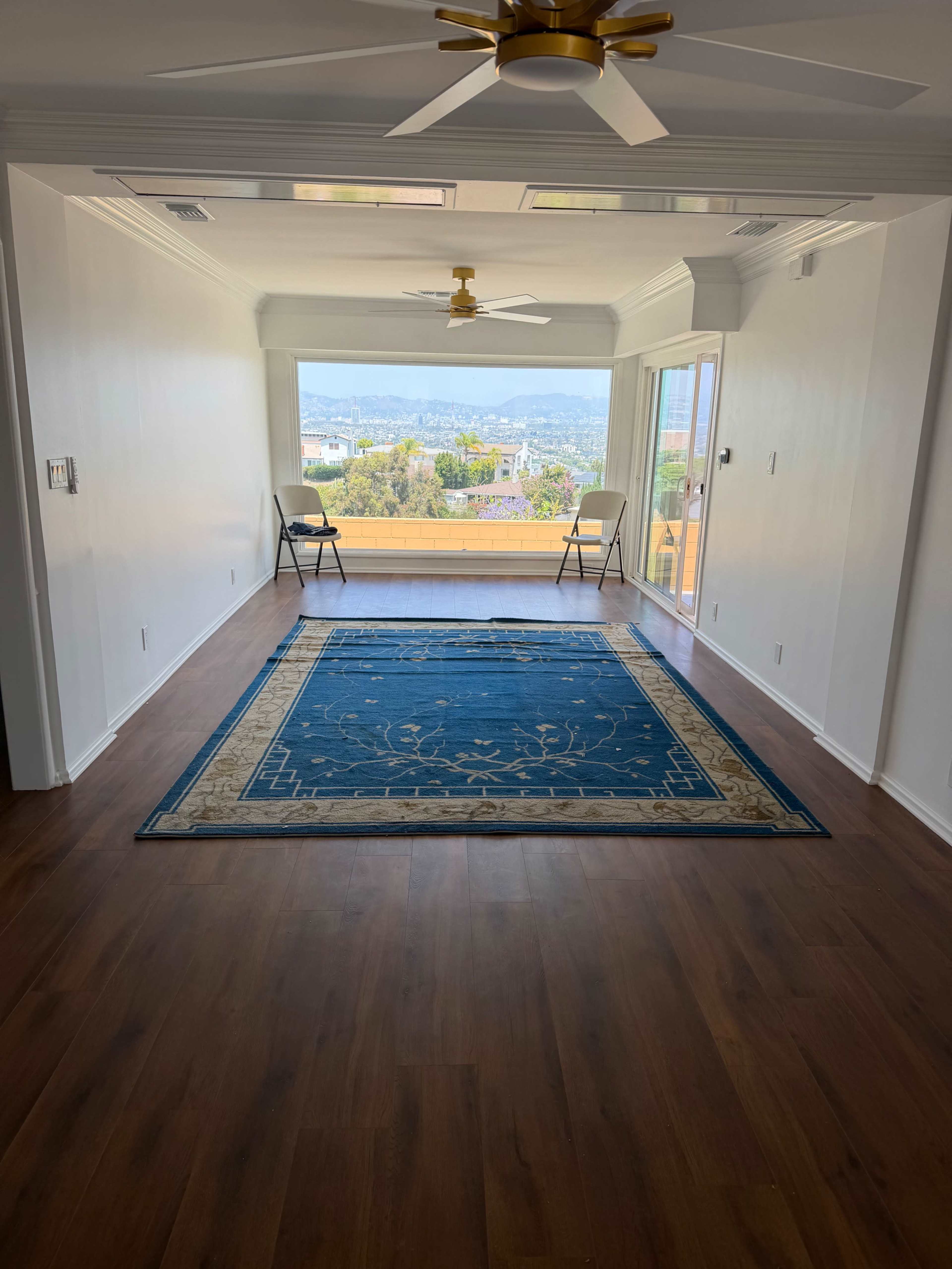 The image shows a brightly lit, empty room with a blue area rug, two folding chairs, and large windows overlooking a scenic view.