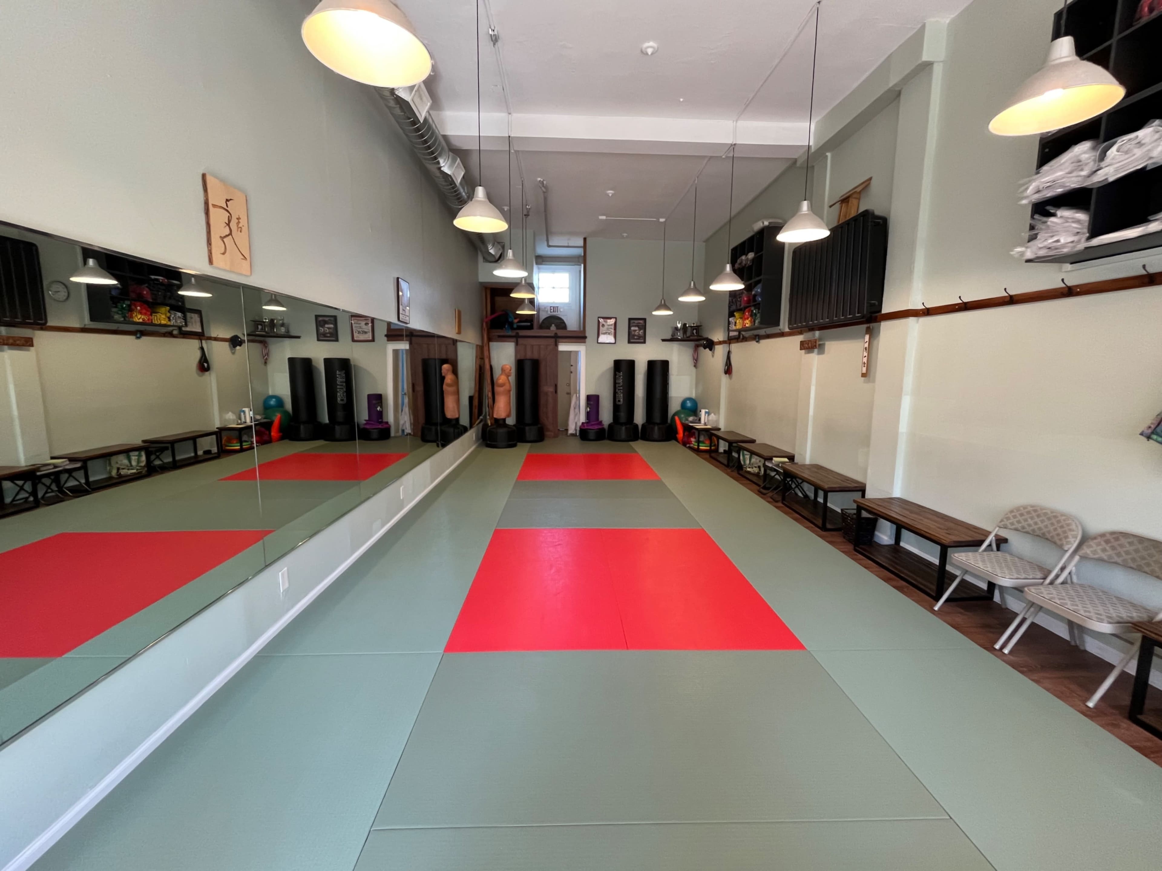 The image shows an empty martial arts studio with mats in the center, mirrors on one wall, and benches along the side.