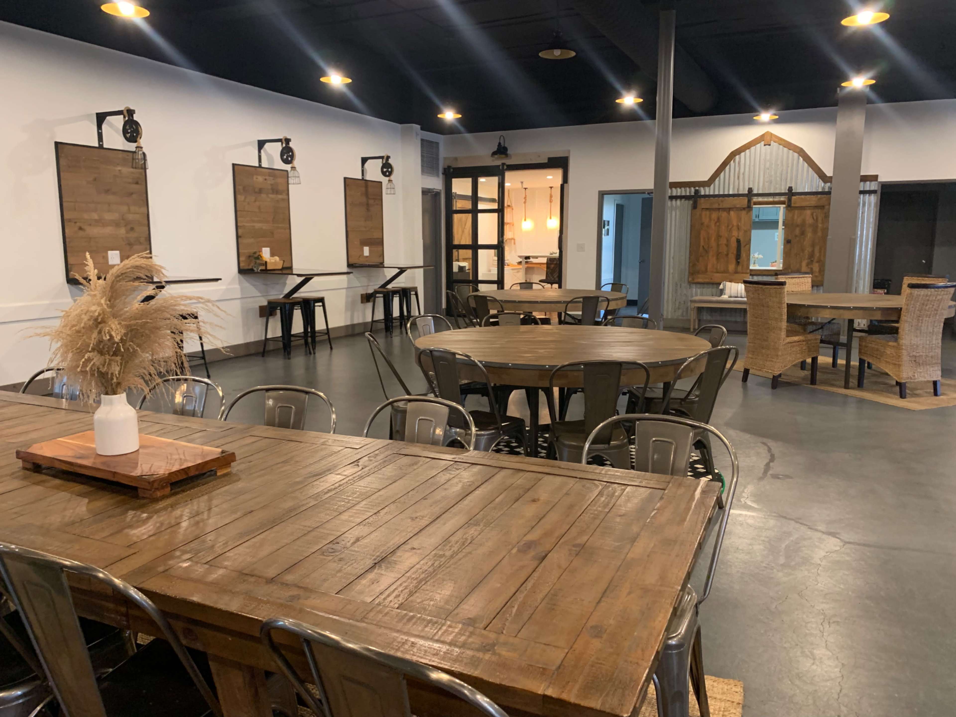 The image shows a spacious interior with multiple wooden tables and chairs, featuring a rustic decor style, lamps, and a cozy seating area in the back.