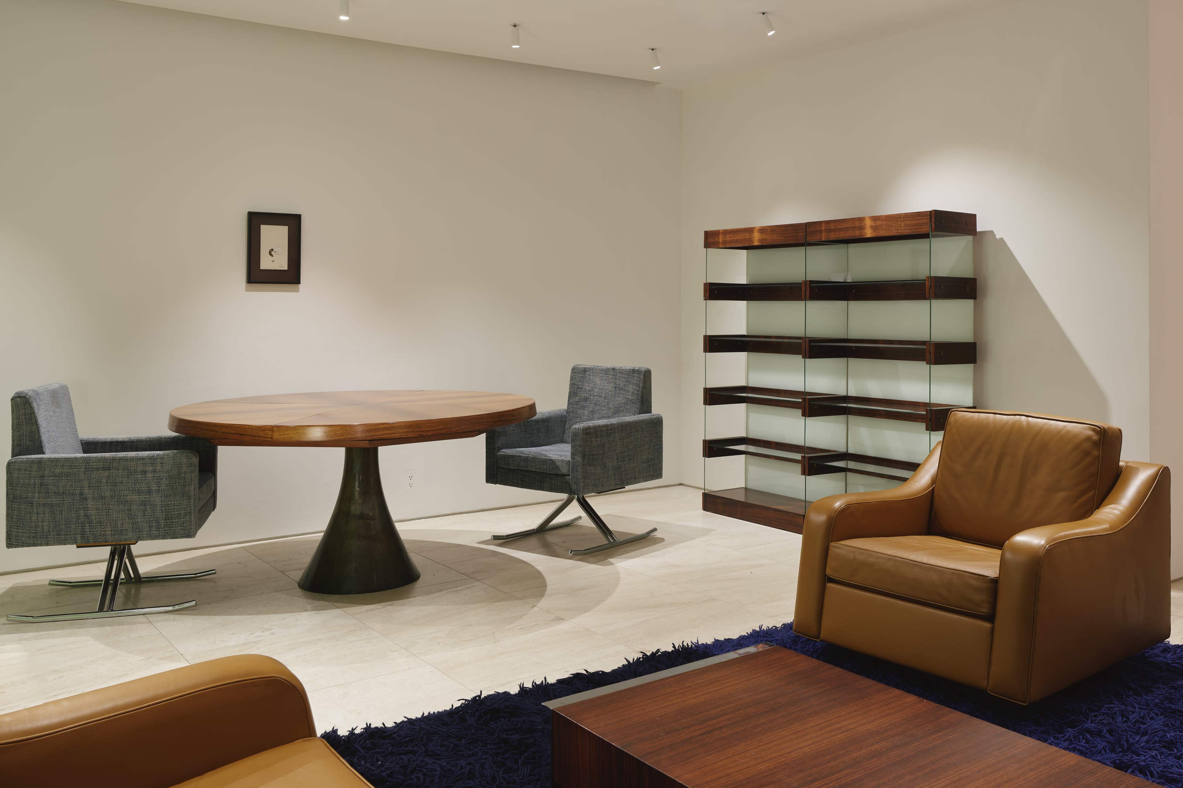 The image shows a modern, minimalistic room featuring a round wooden table with two grey chairs, a brown leather armchair, and a glass display cabinet against a white wall.