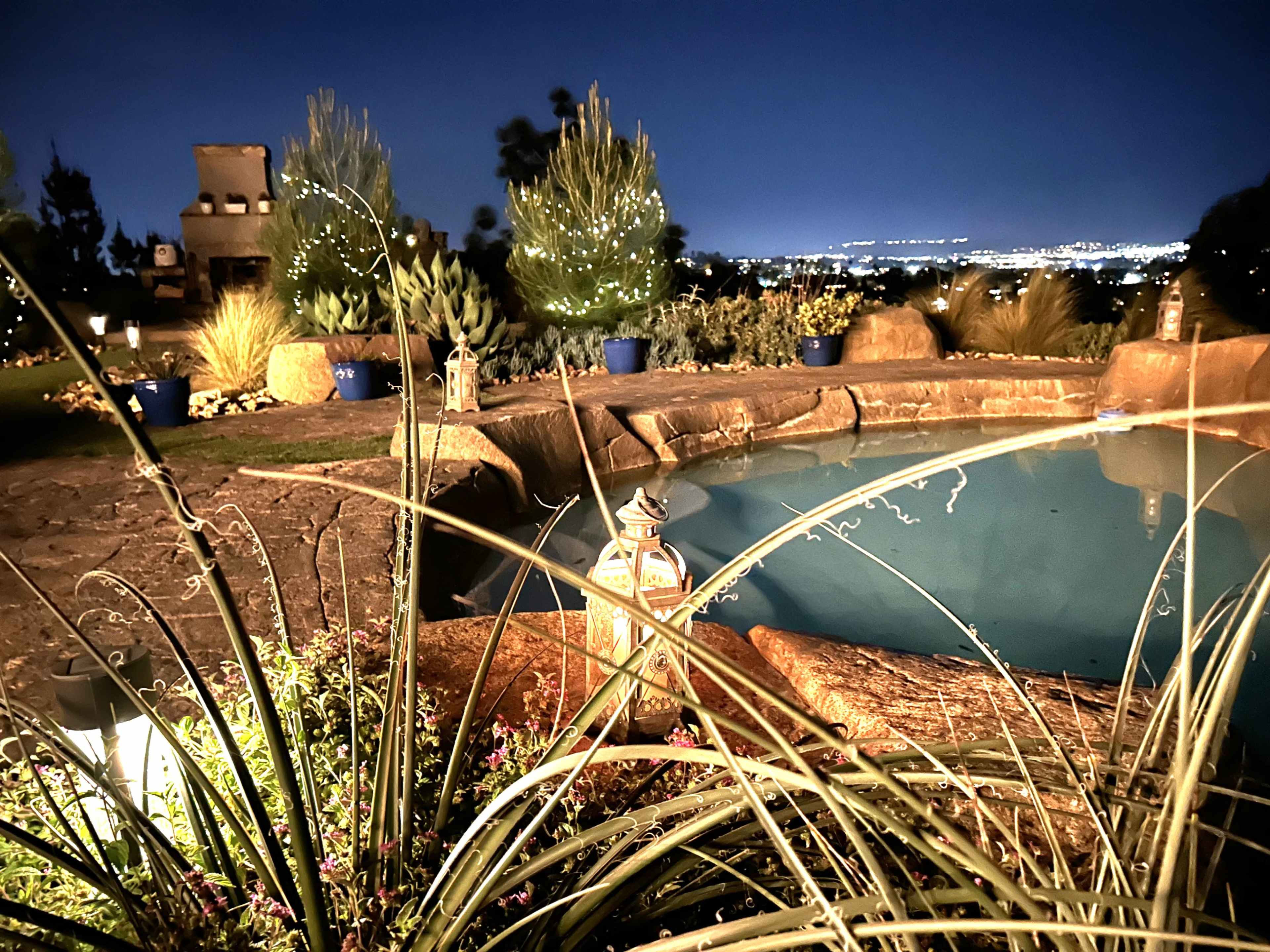 A illuminated backyard pool area at night, surrounded by greenery and decorative lanterns.