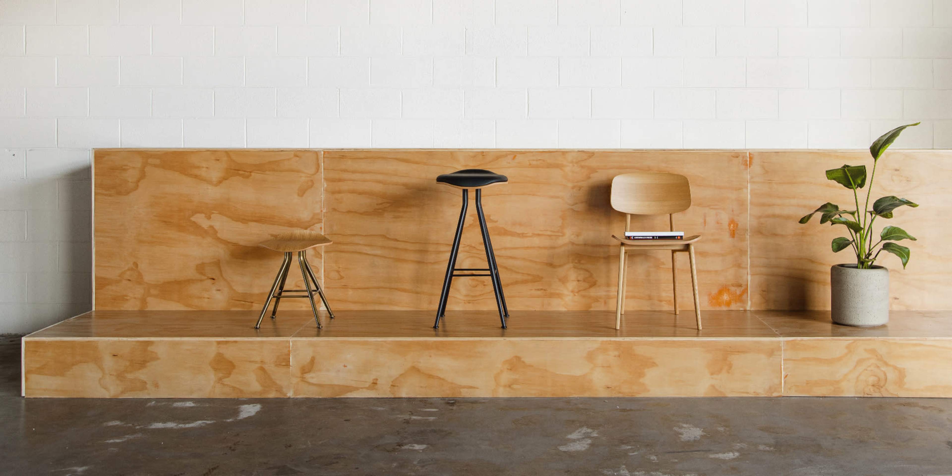 A simple wooden platform with three stools of different designs and a potted plant against a white wall.