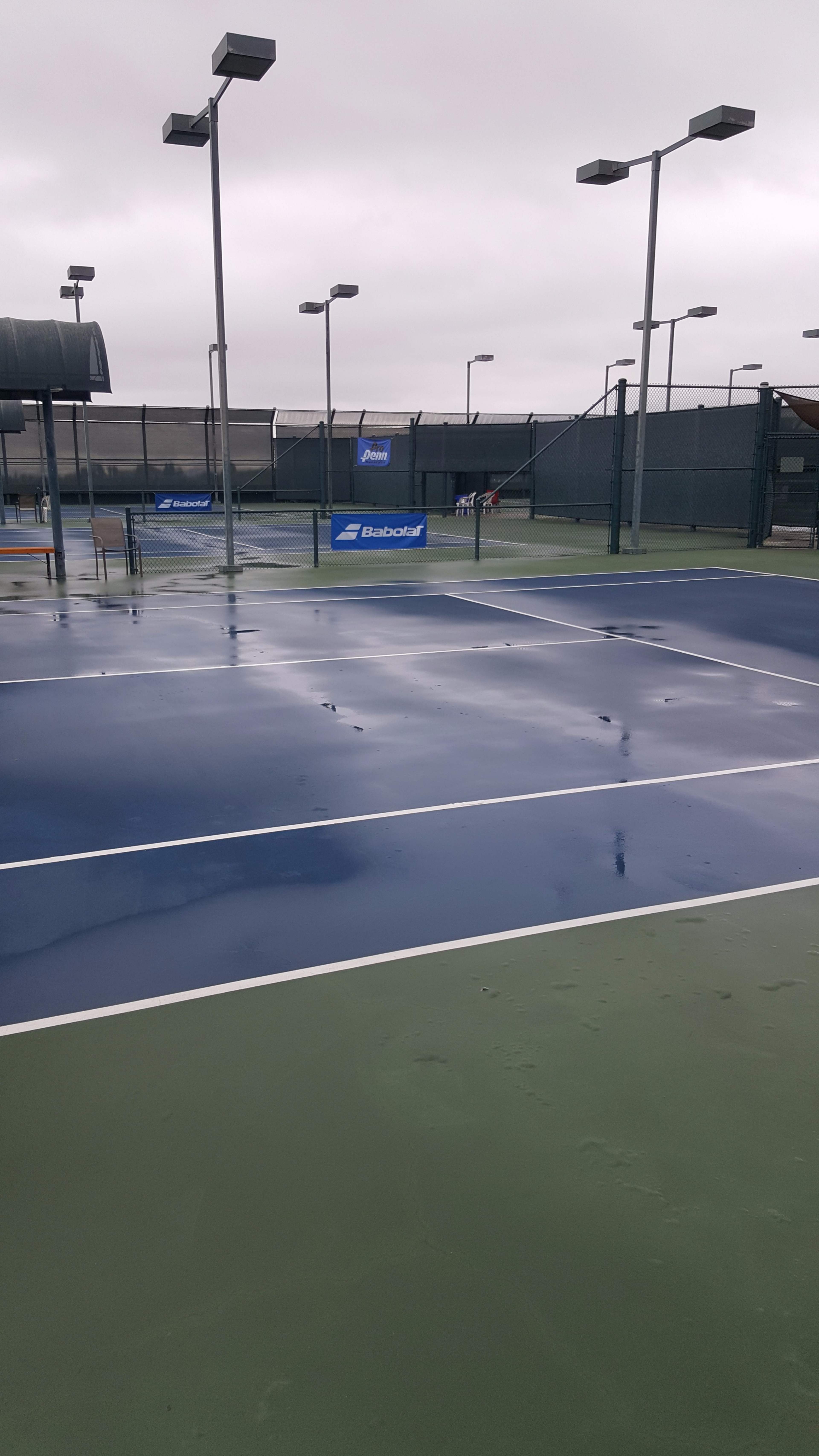 A set of wet tennis courts with blue and green surfaces, surrounded by poles with lights and a covered structure in the background.