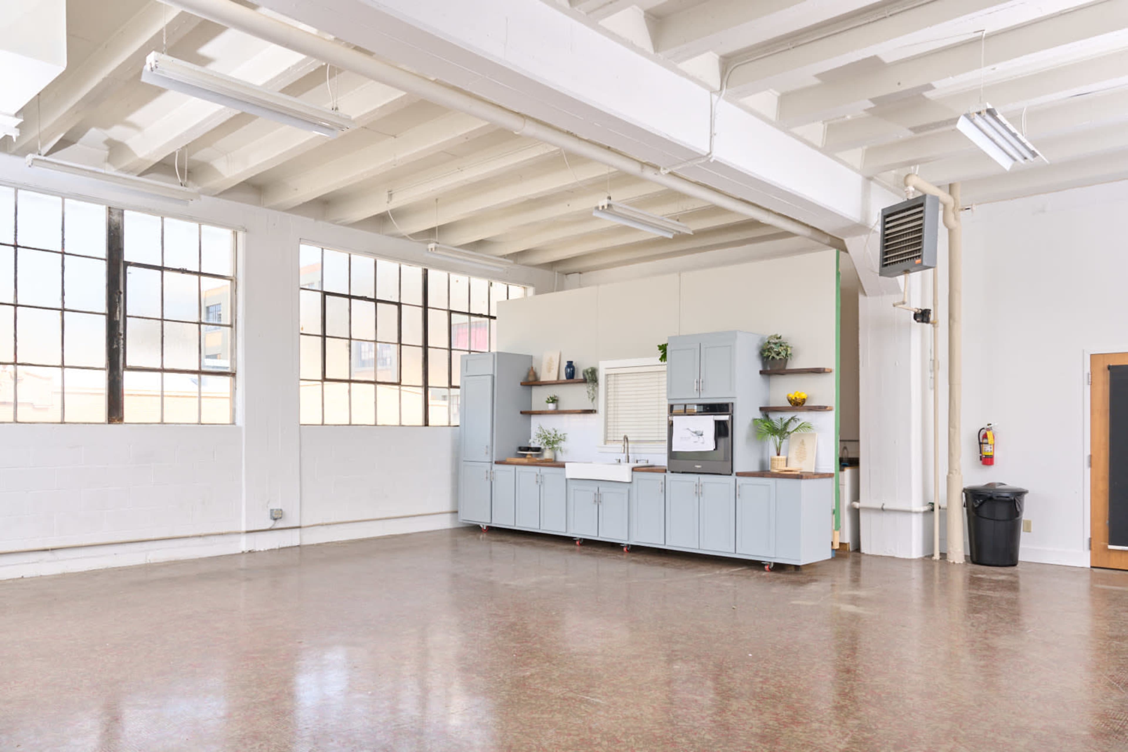 The image shows a spacious room with a kitchen area featuring light blue cabinets and large windows allowing natural light to enter.