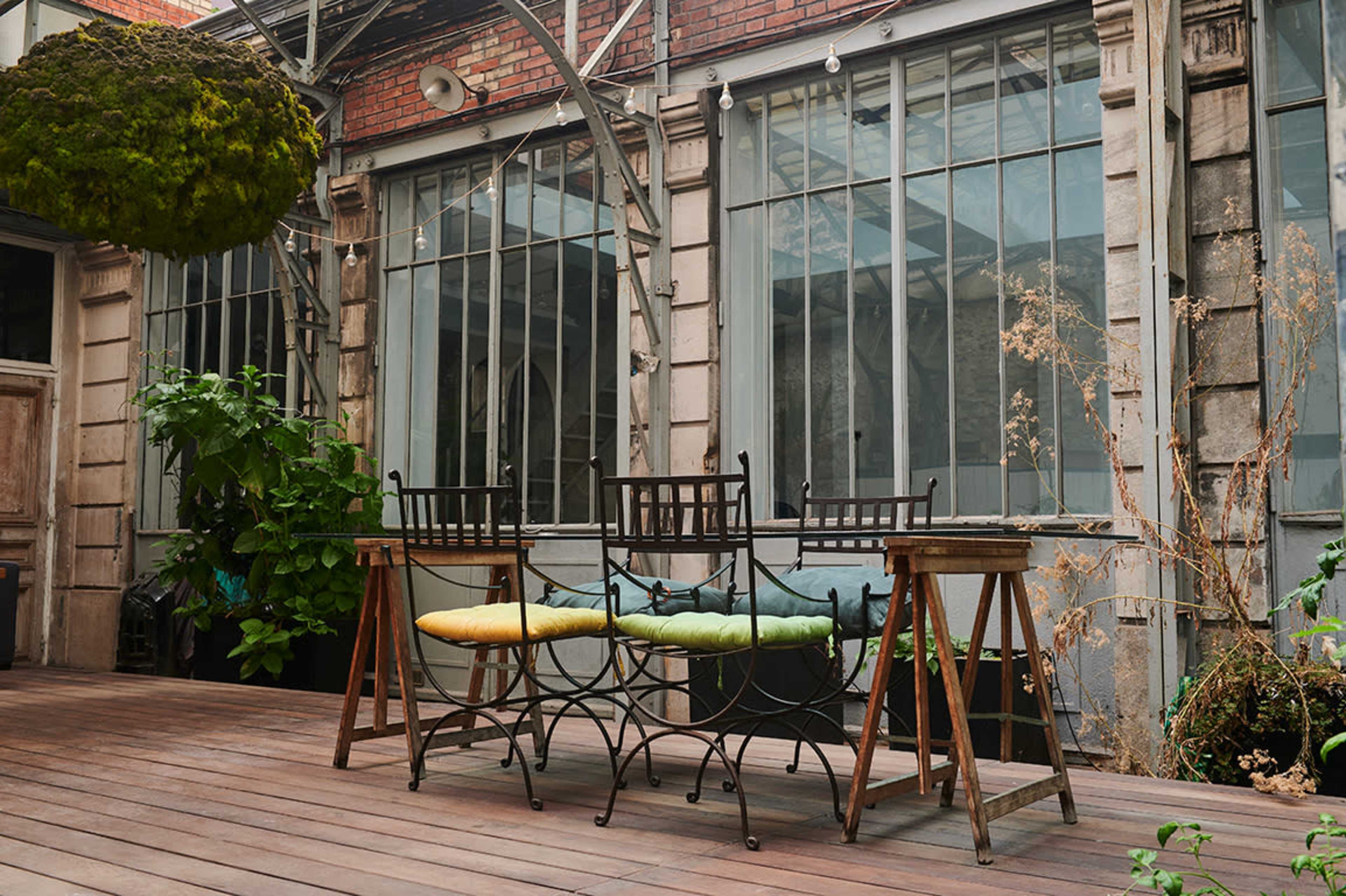 A wooden deck features a metal table with four chairs, surrounded by plants and large windows in a rustic building.