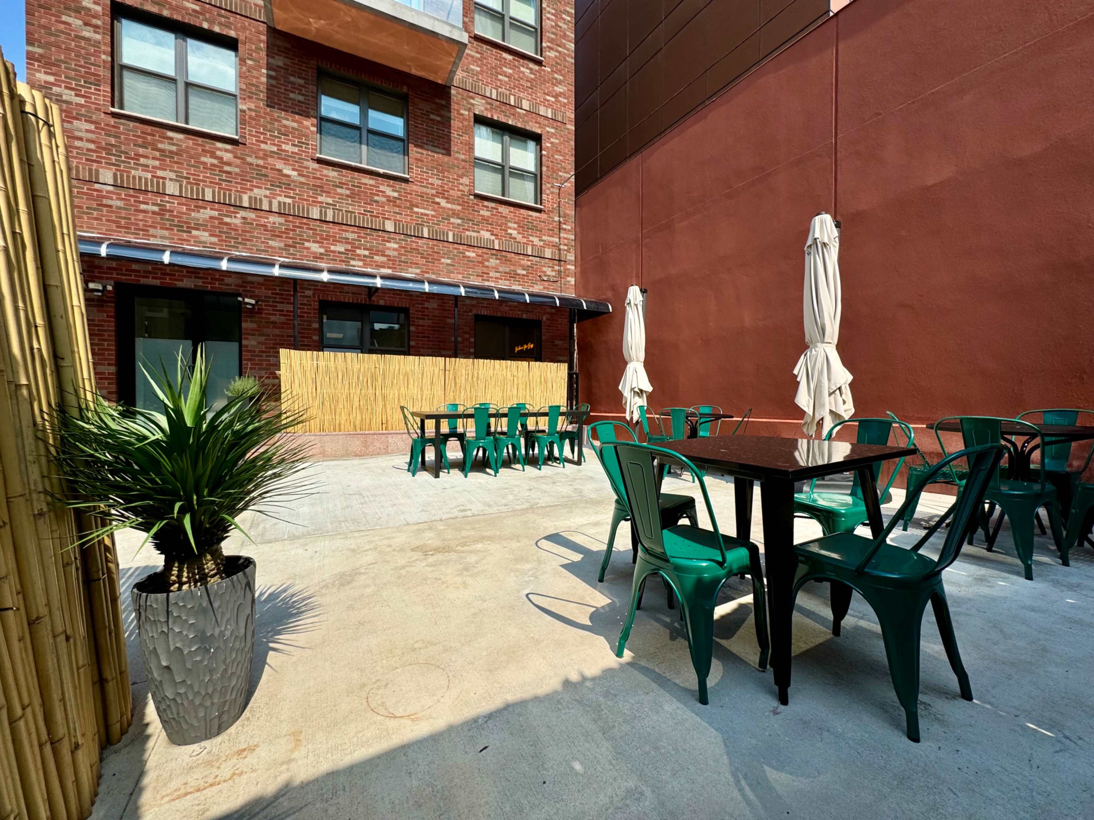 An outdoor seating area with green metal chairs and tables, surrounded by a brick building and bamboo screens.