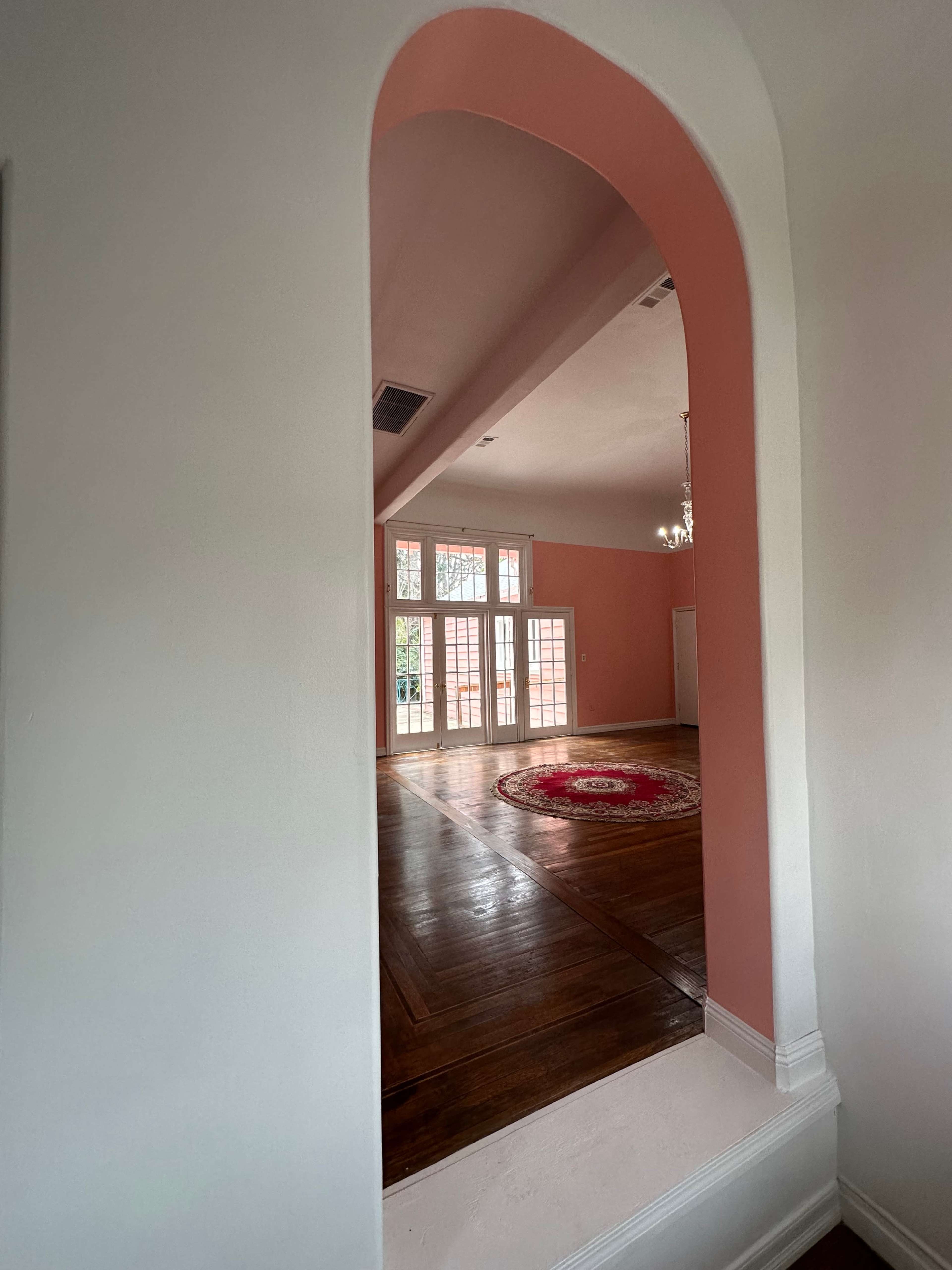 A view through an arched opening reveals a spacious room with a chandelier and large windows, featuring a round rug on the wooden floor.