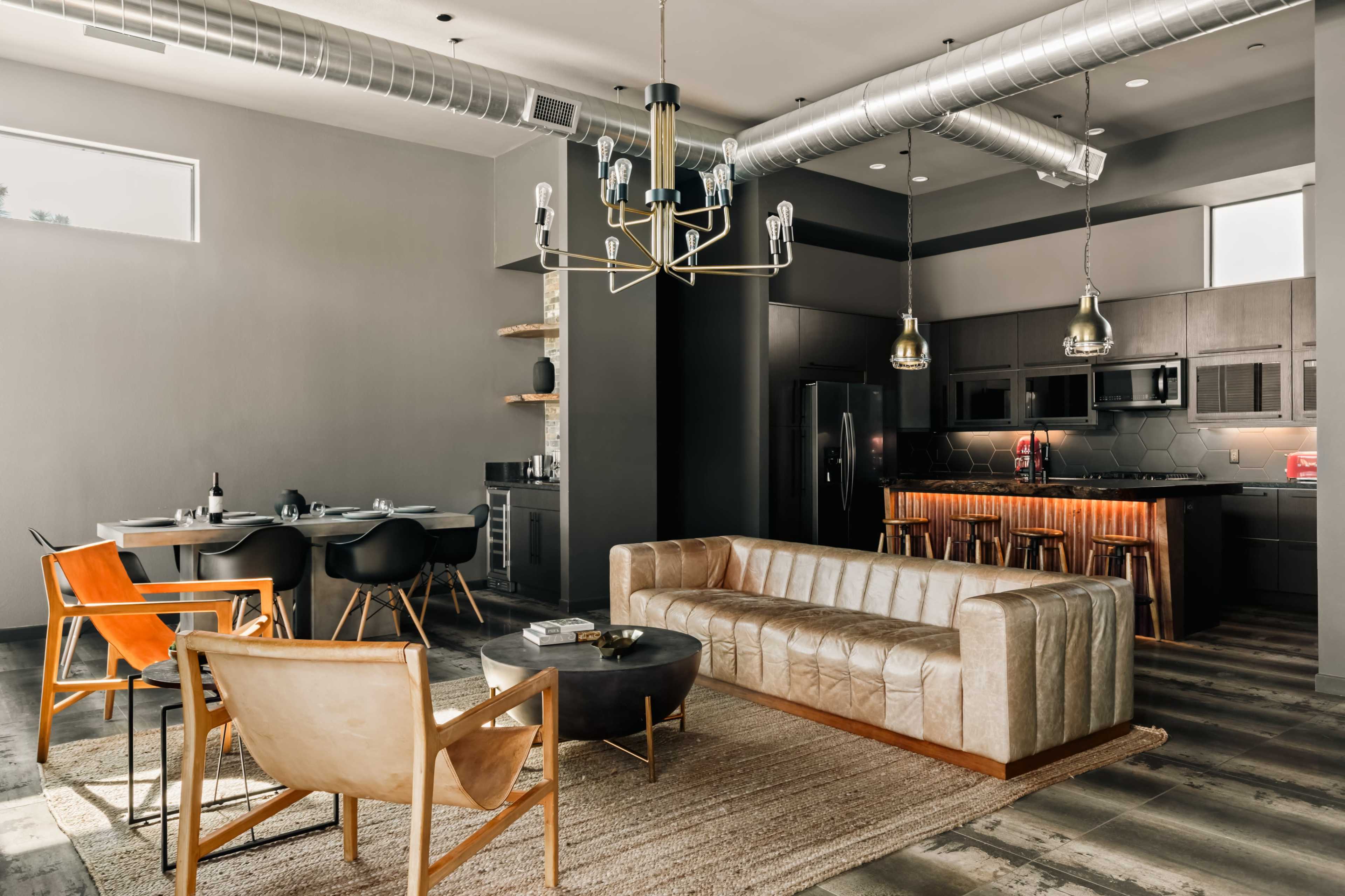 The image shows a modern living room with a beige tufted sofa, two armchairs, a round coffee table, and a dining area in the background, all within a stylish industrial space.