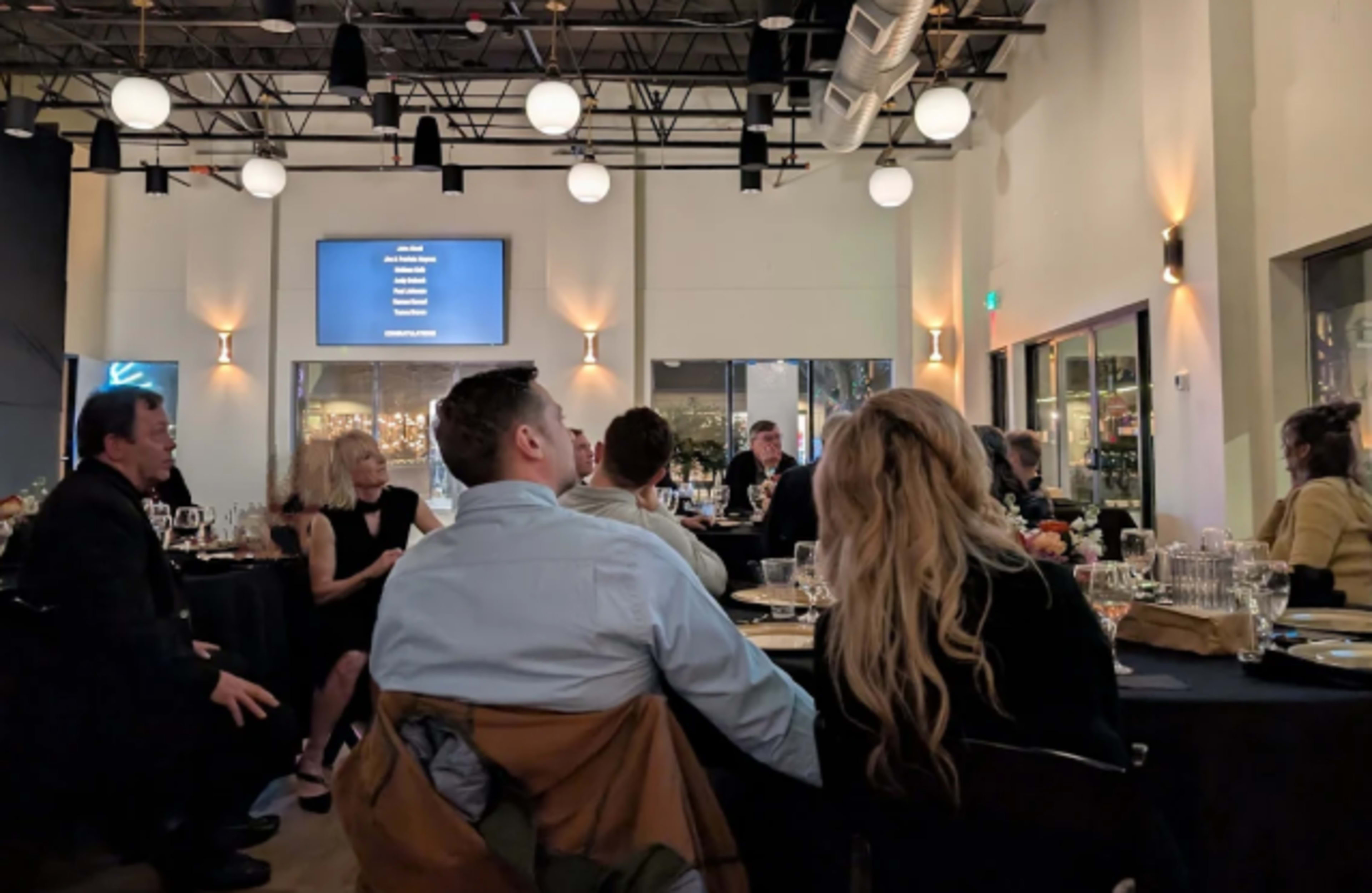 A group of people sits around tables in a well-lit dining area, paying attention to a screen displaying information or a presentation.