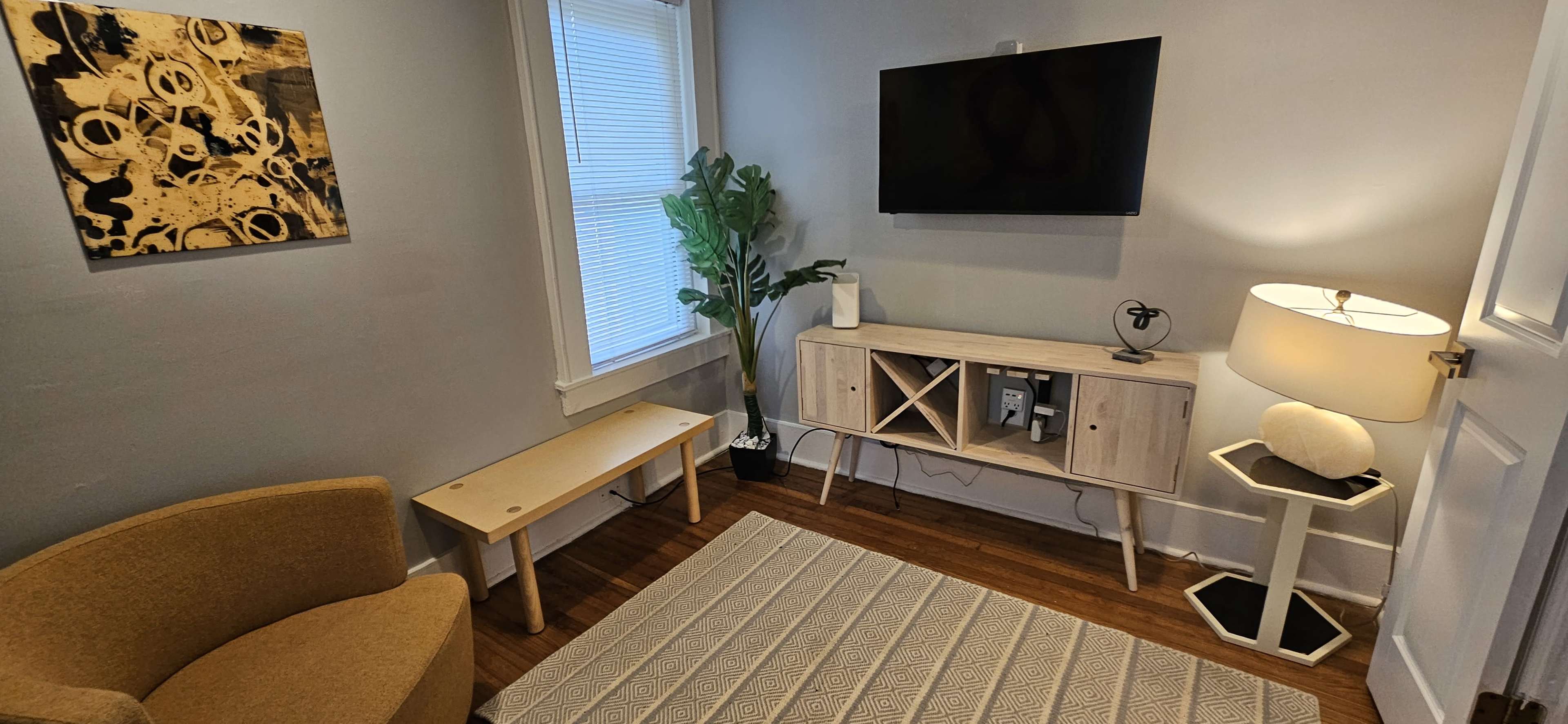 A small living area features a beige chair, a wooden bench, a television mounted on the wall, a light-colored media console, a potted plant, and a lamp.