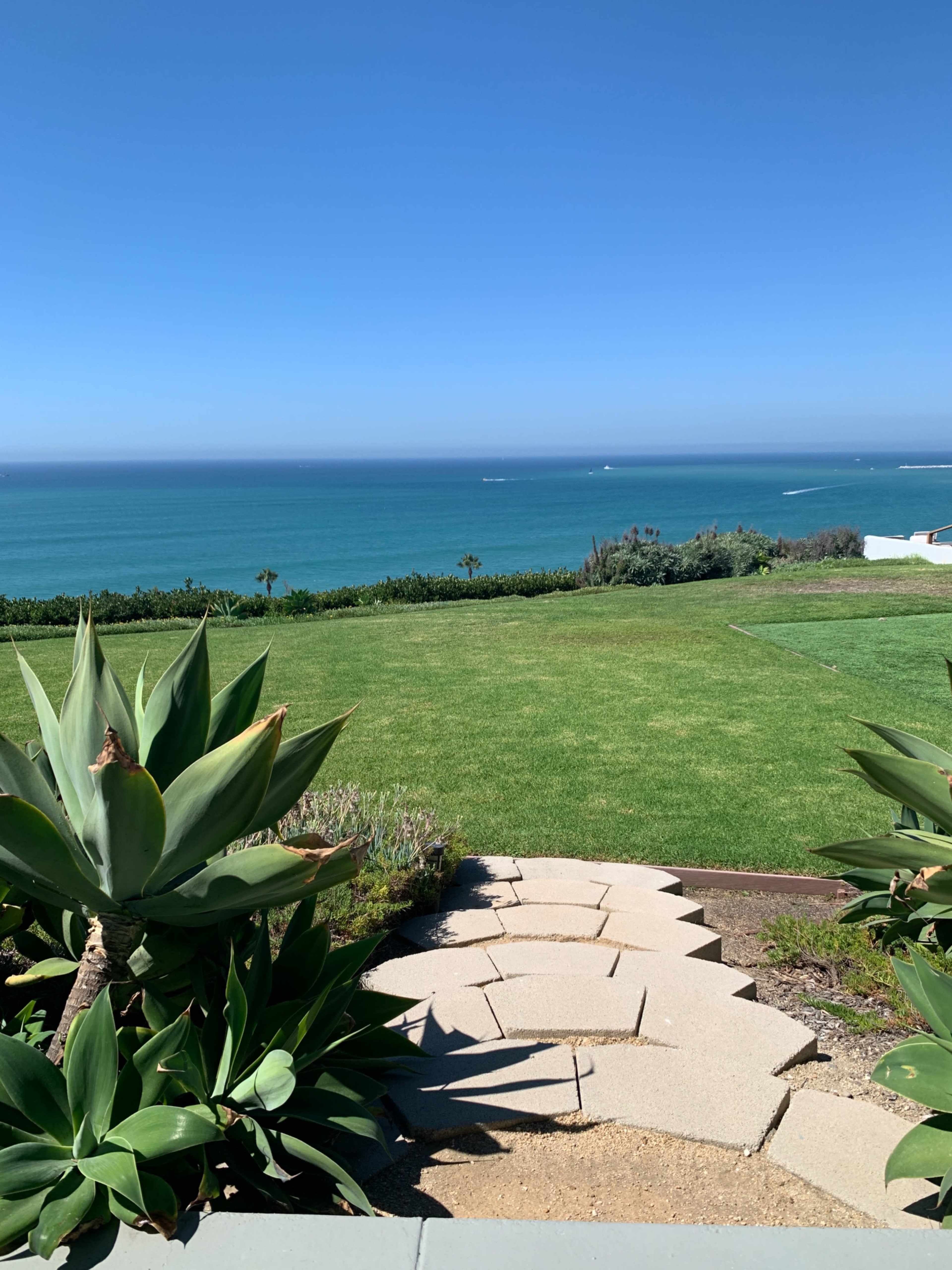 A pathway of stone steps leads down to a grassy area overlooking the blue ocean and clear sky.