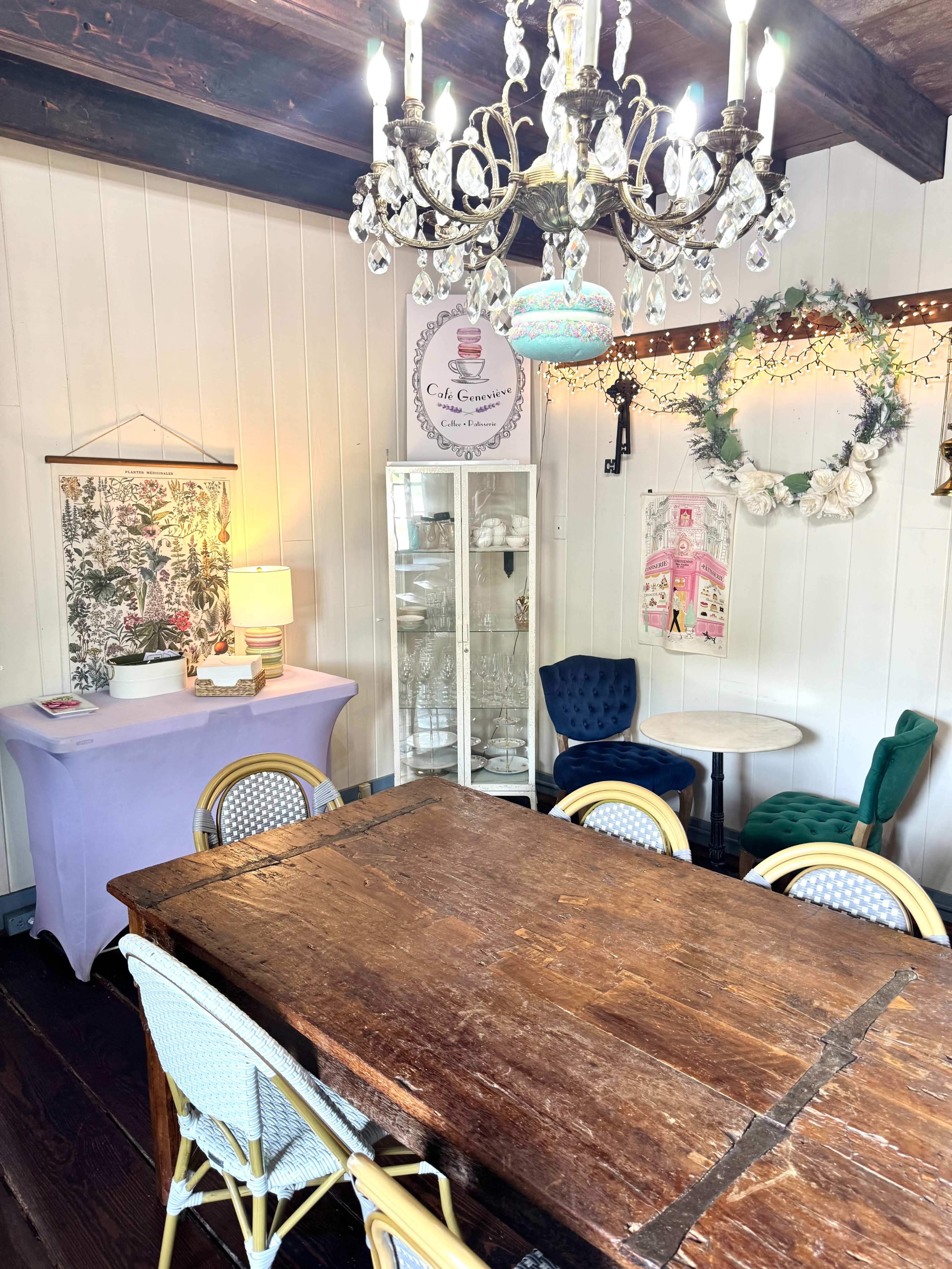 The image shows a cozy room featuring a wooden dining table surrounded by light-colored chairs, a chandelier overhead, and decorative elements on the walls.