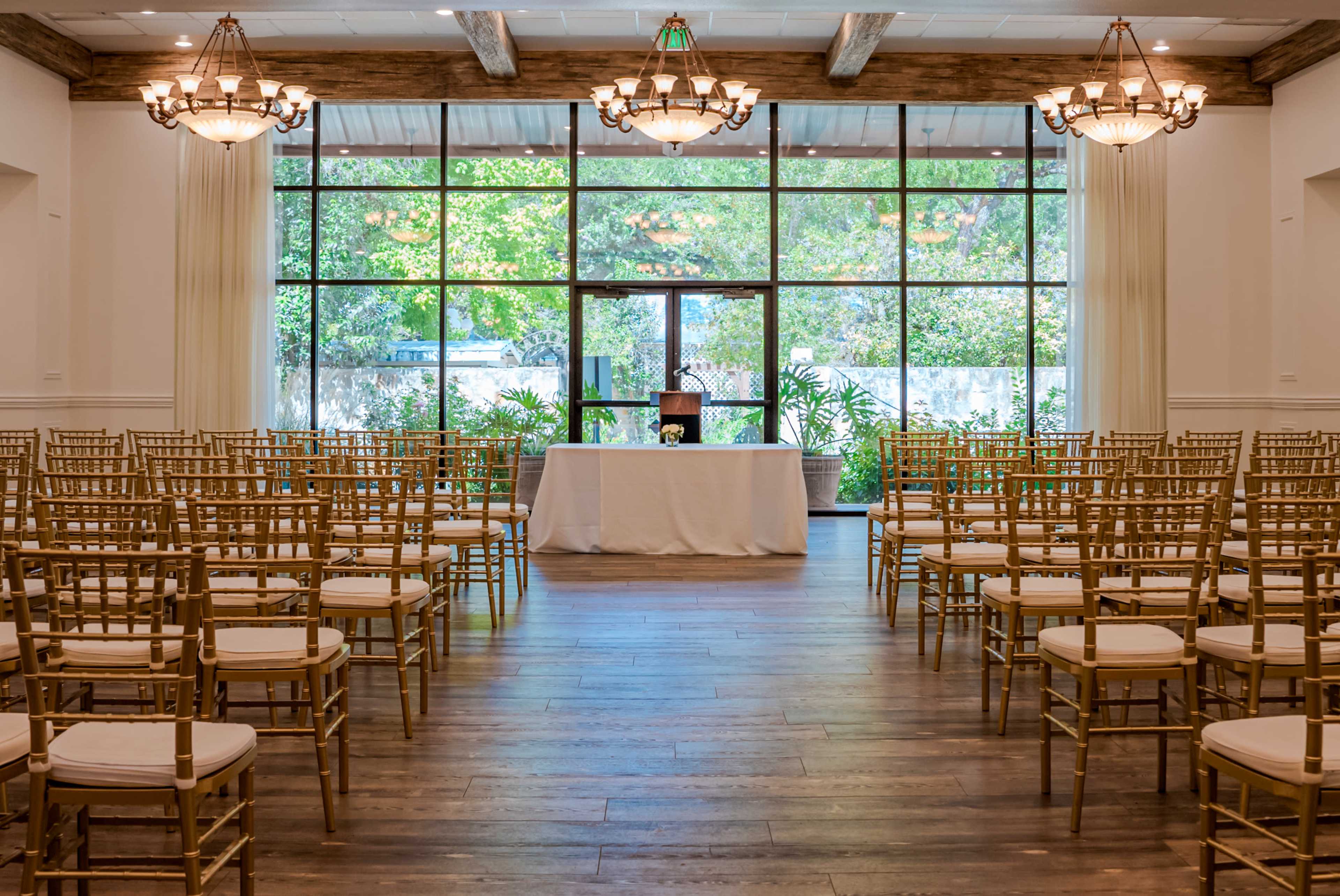 The image shows a ceremony space arranged with rows of gold chiavari chairs facing a small altar under large windows that let in natural light.
