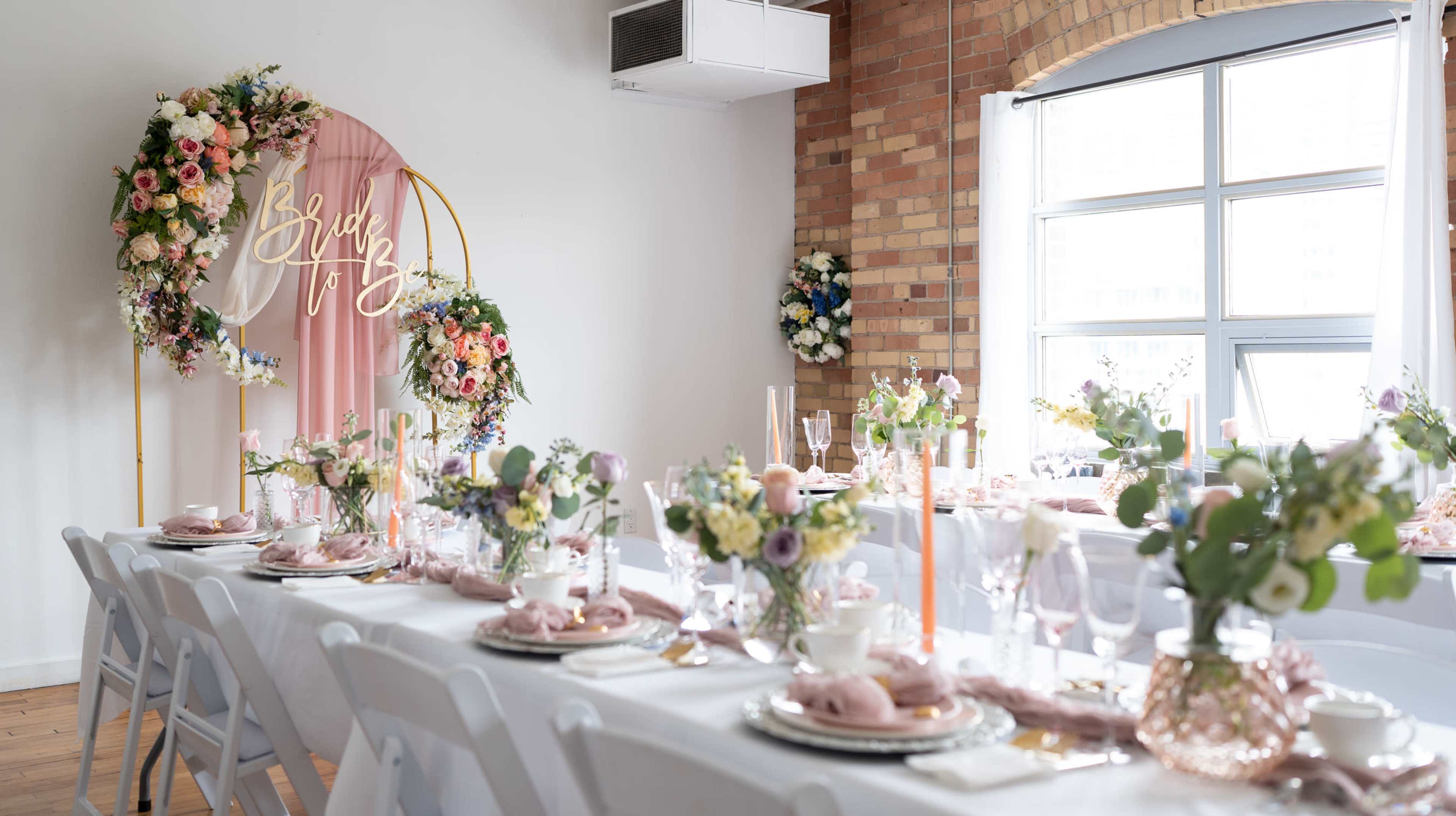 A beautifully arranged dining table is set with white chairs, floral centerpieces, and elegant tableware in a warmly decorated room featuring a floral arch.
