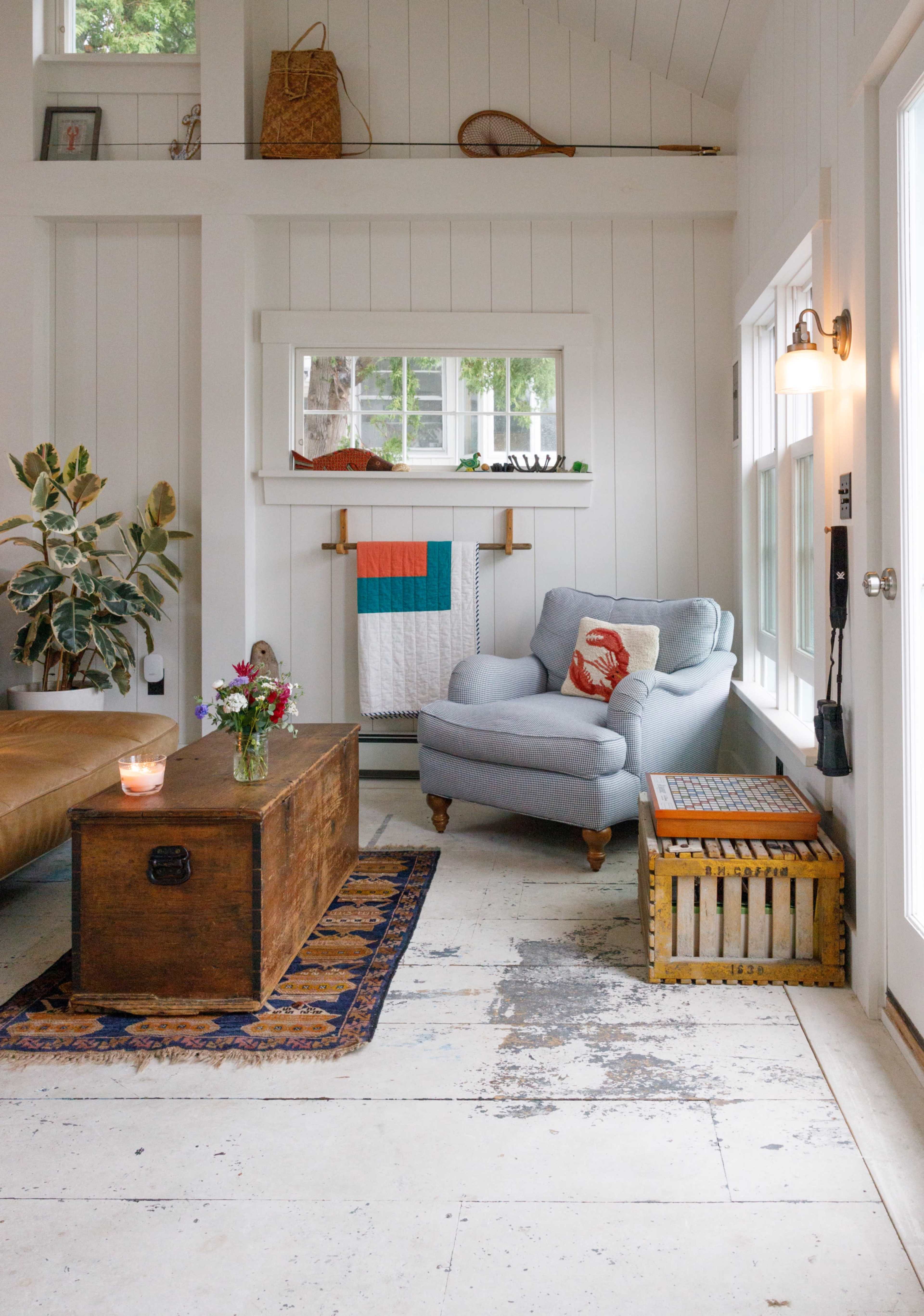The image shows a cozy living area featuring a light blue armchair, a brown coffee table, and a potted plant, with white walls and large windows letting in natural light.