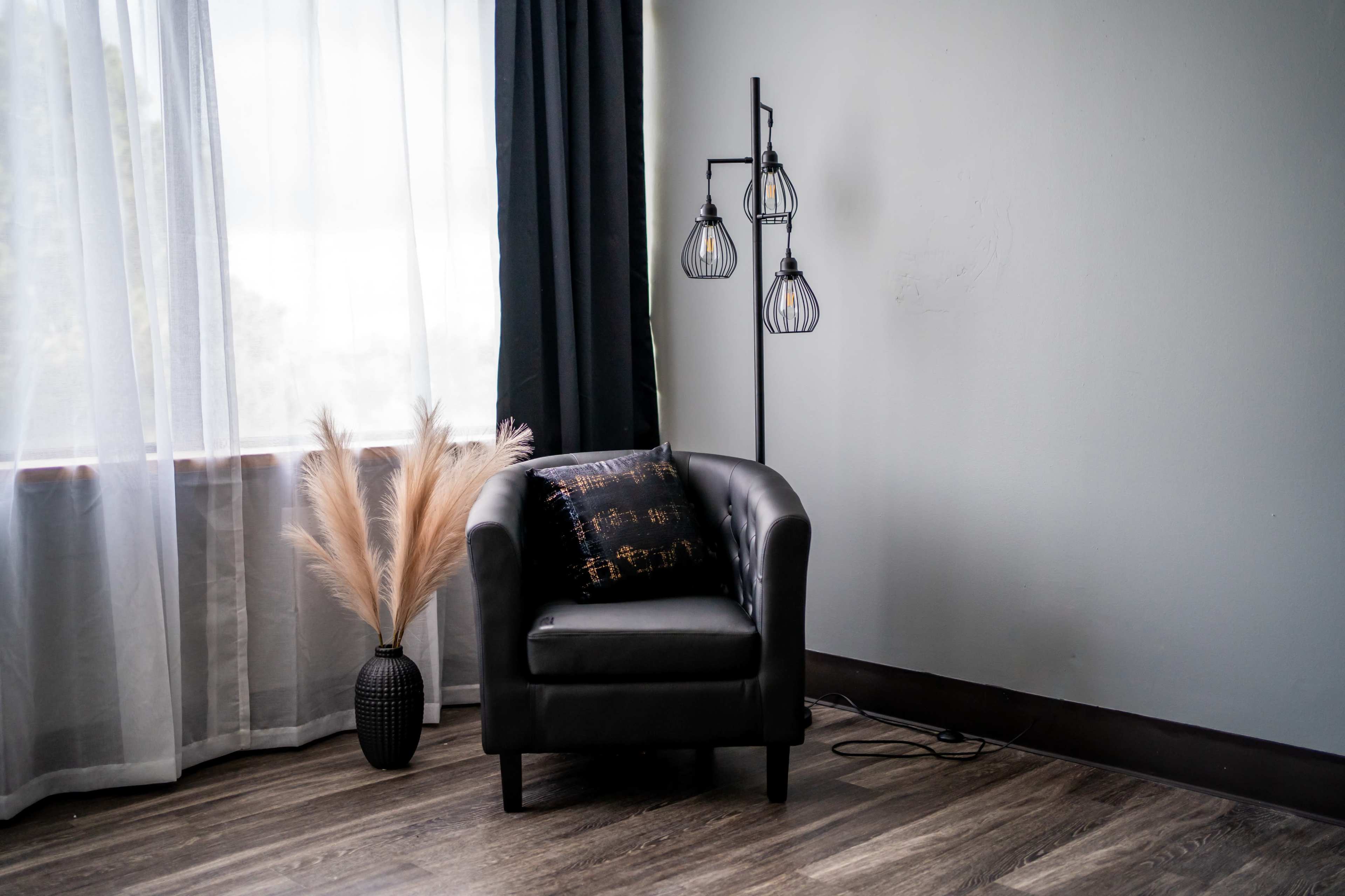 A black armchair sits next to a floor lamp with three exposed bulbs, beside a tall vase containing dried grasses, in a softly lit room with sheer white curtains.