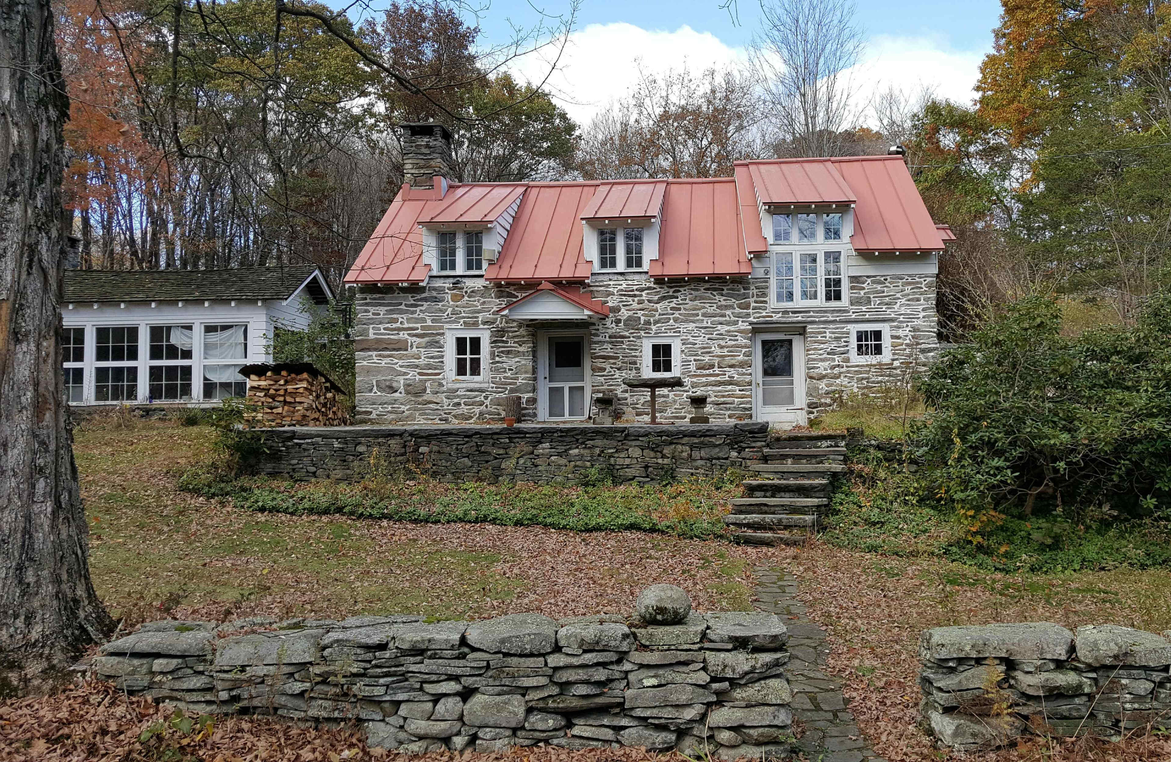 A stone cottage with a red metal roof surrounded by trees and autumn foliage, with a pathway leading to its entrance.