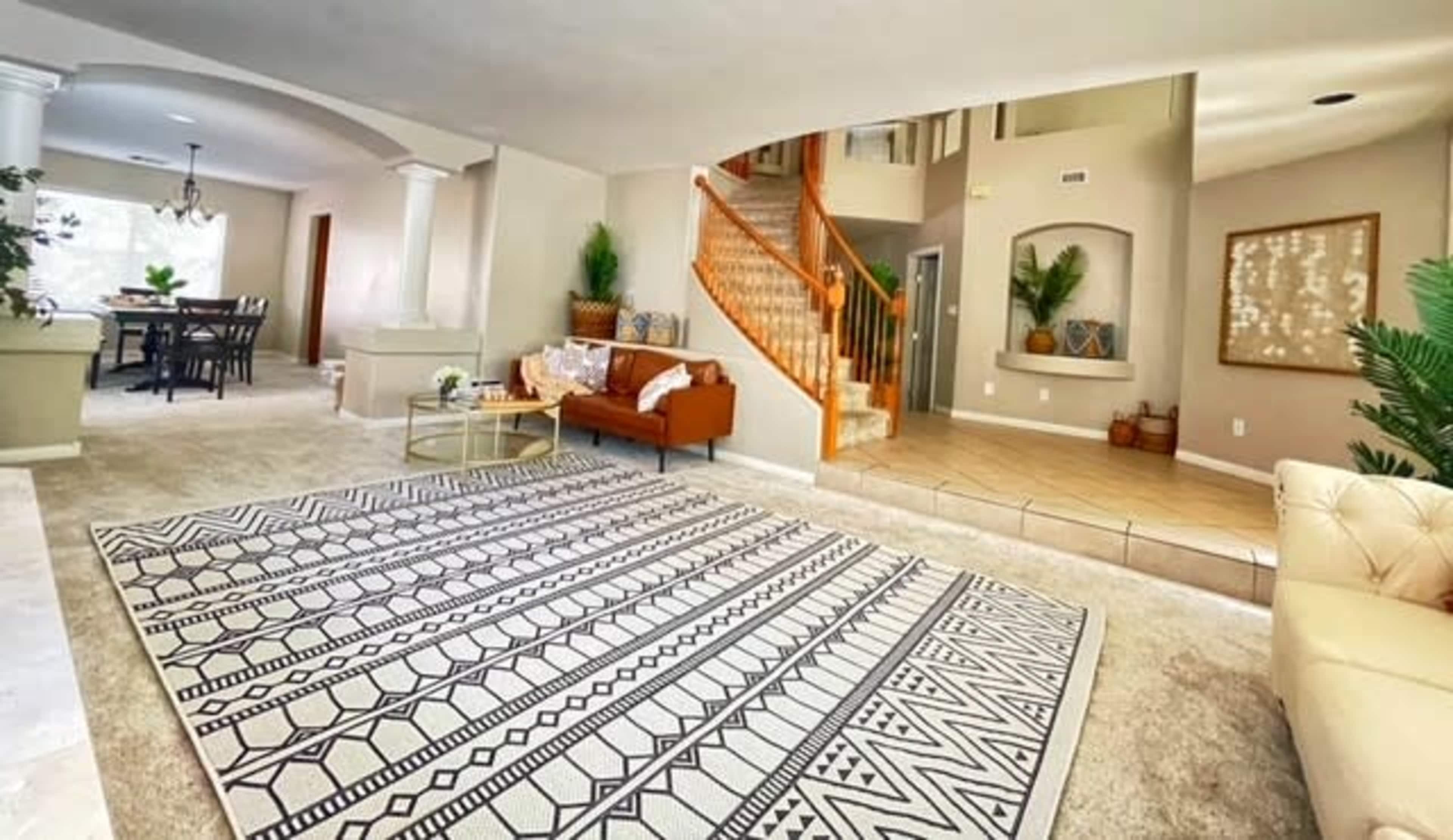 A spacious living room with beige carpeting, a patterned area rug, a brown sofa, and a staircase leading to an upper floor.