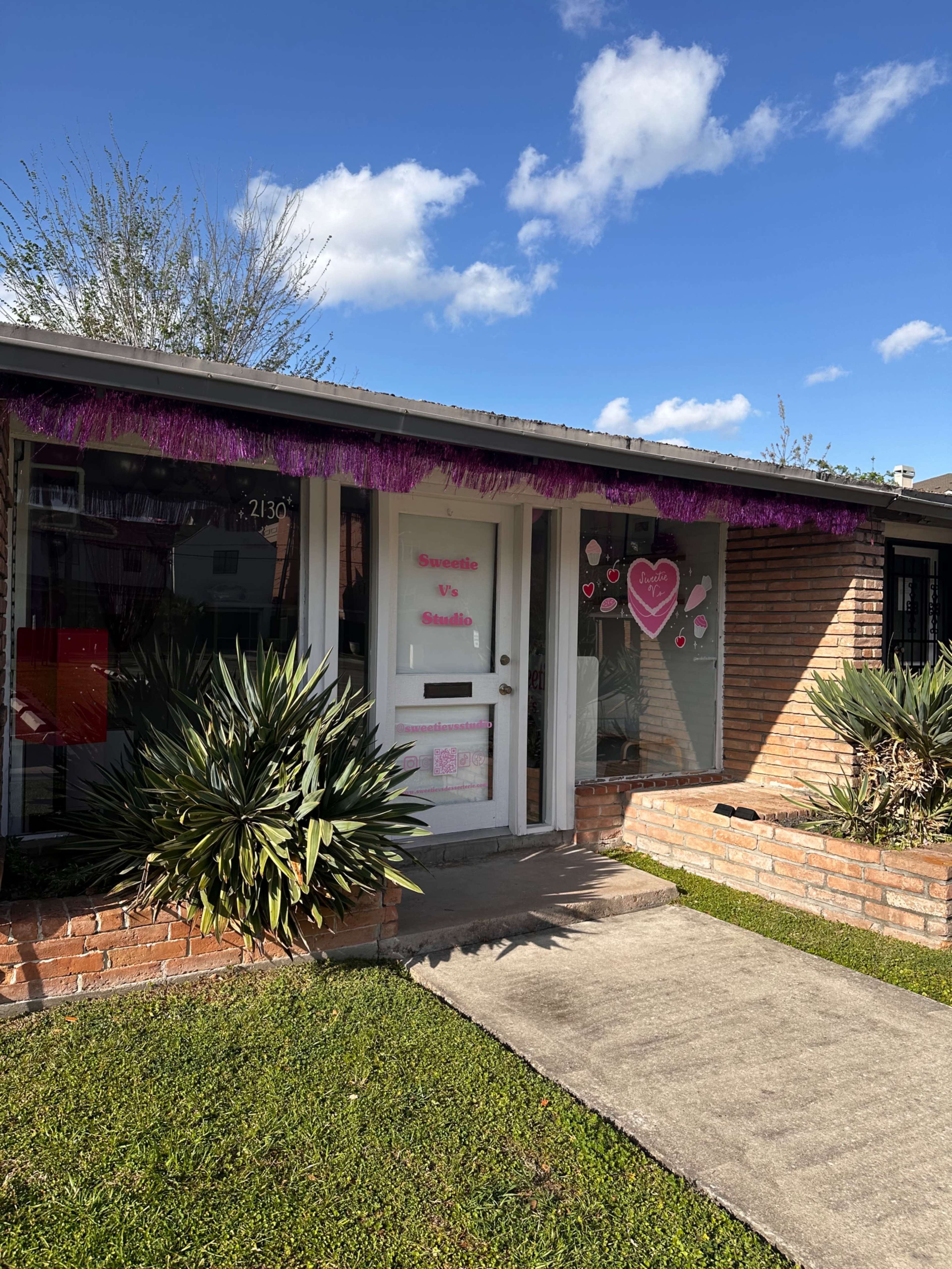 The image shows a small brick building with a front door adorned with decorations, including hearts and a purple fringe, set against a clear blue sky.