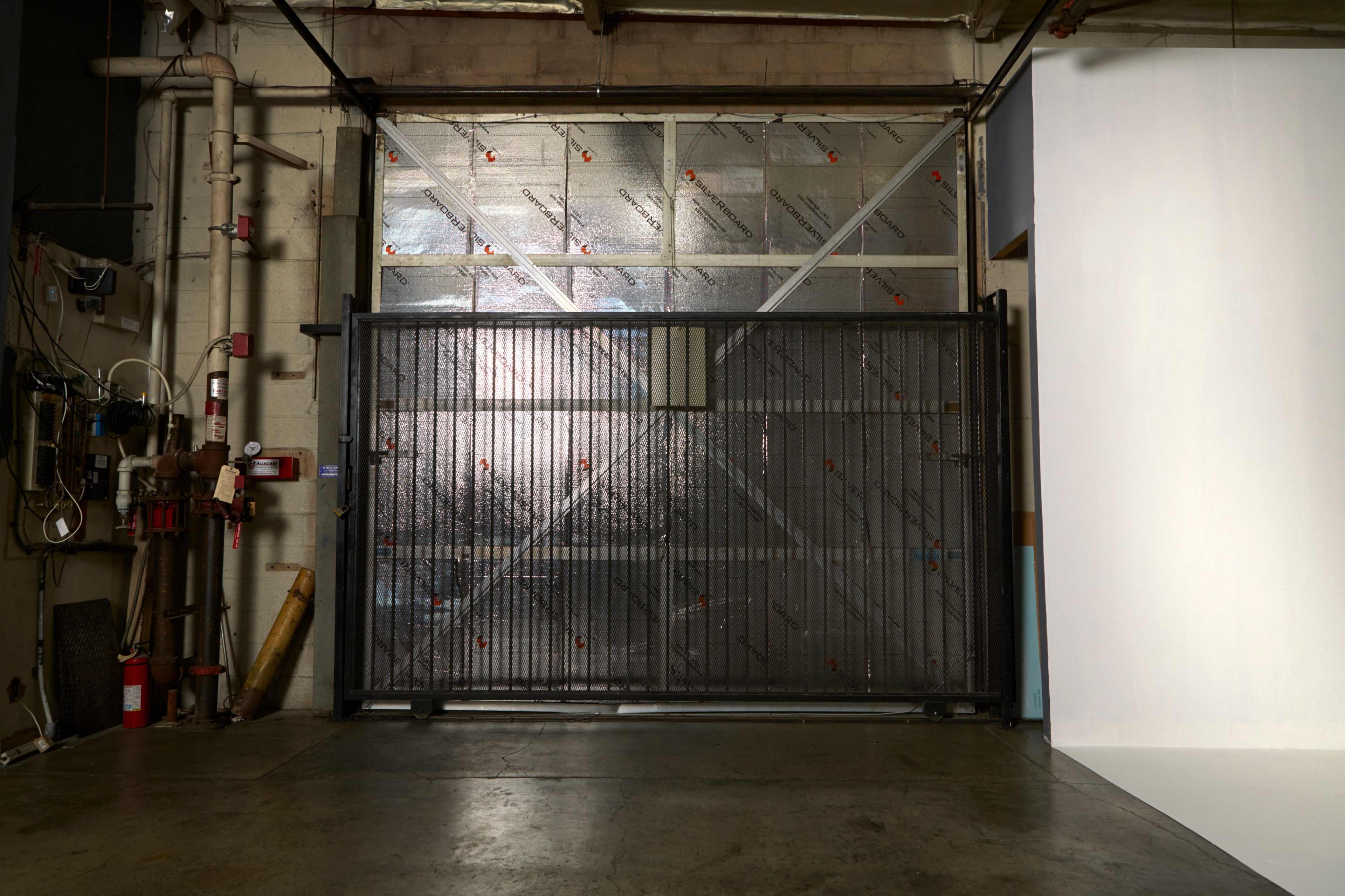 A metal gate installed in a warehouse with a reflective background and exposed pipes on the left side.