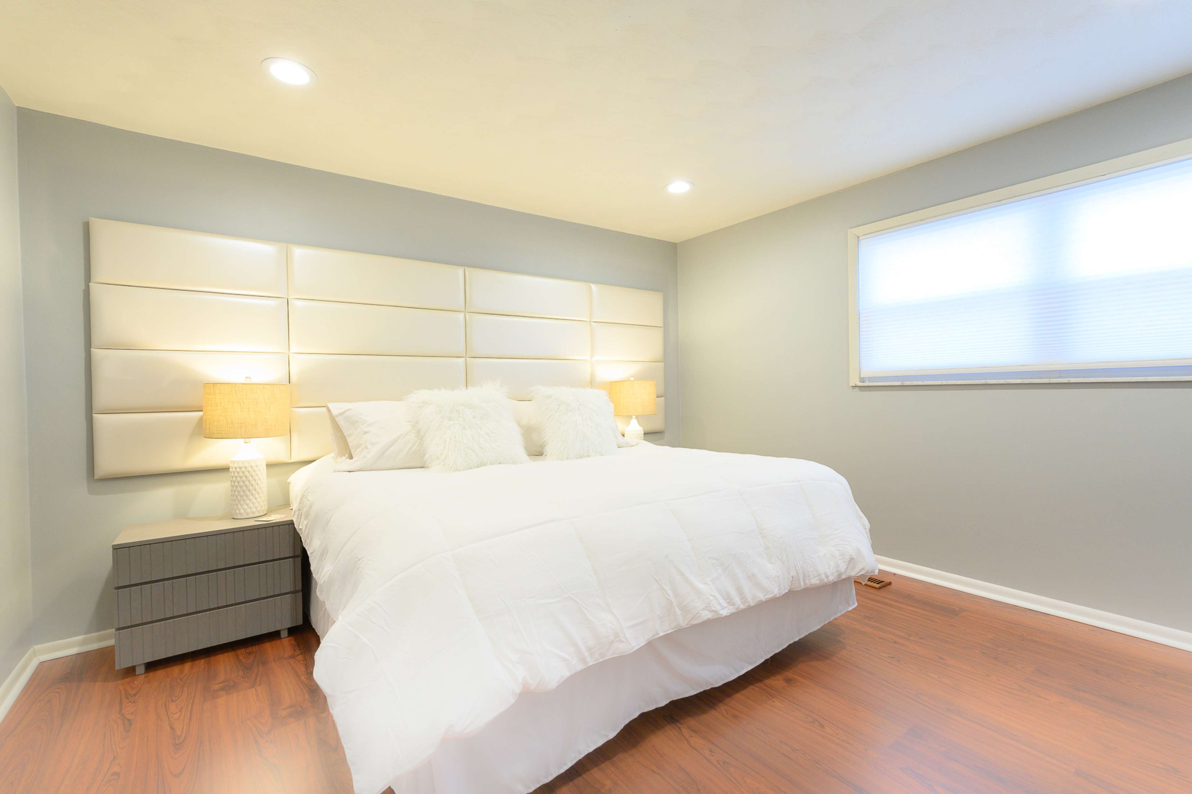 A well-lit bedroom features a large bed with white linens, two bedside tables with lamps, and a large window.