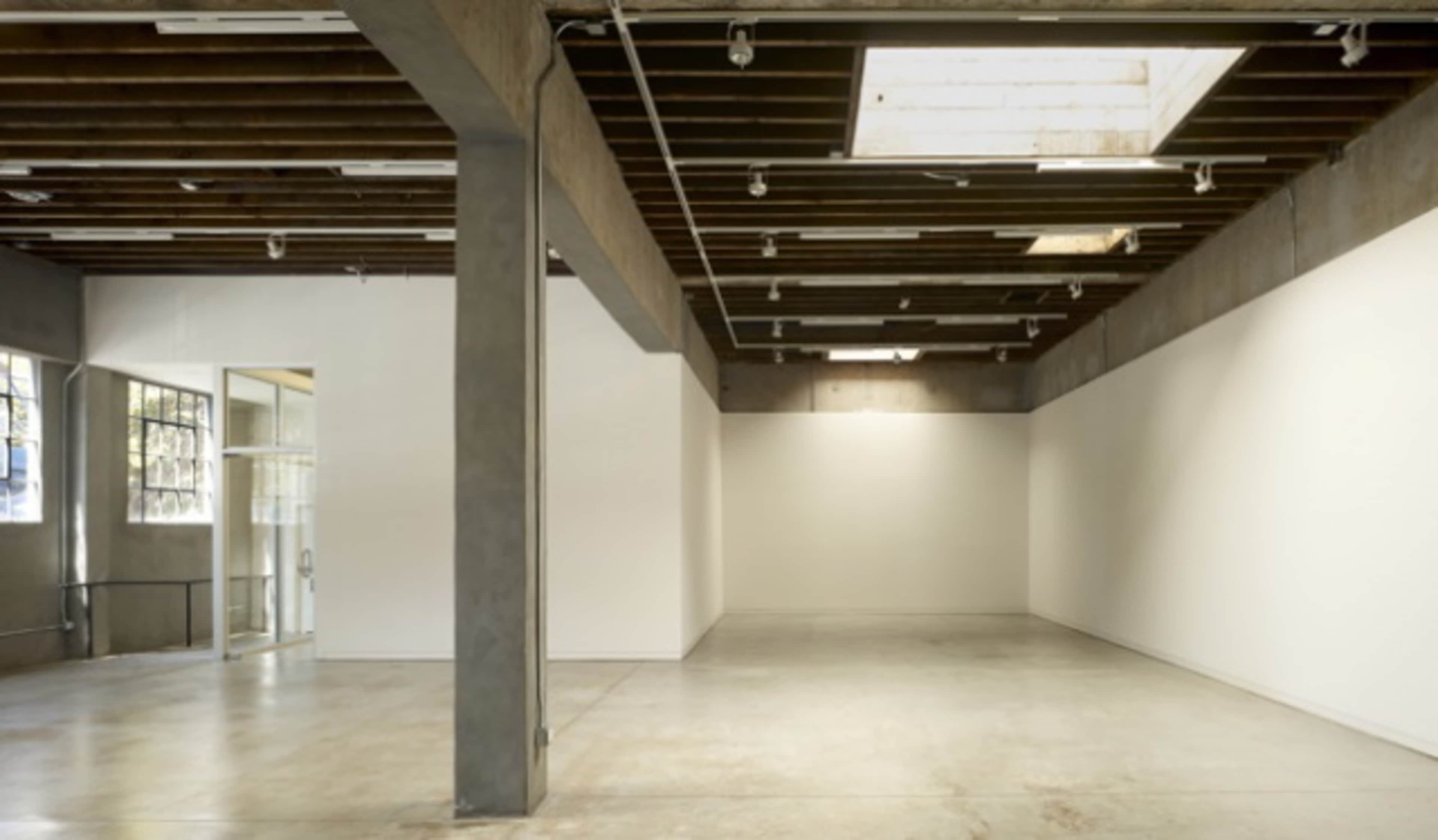 An empty room with concrete walls, exposed wooden beams, and large windows, featuring a polished concrete floor and minimal overhead lighting.