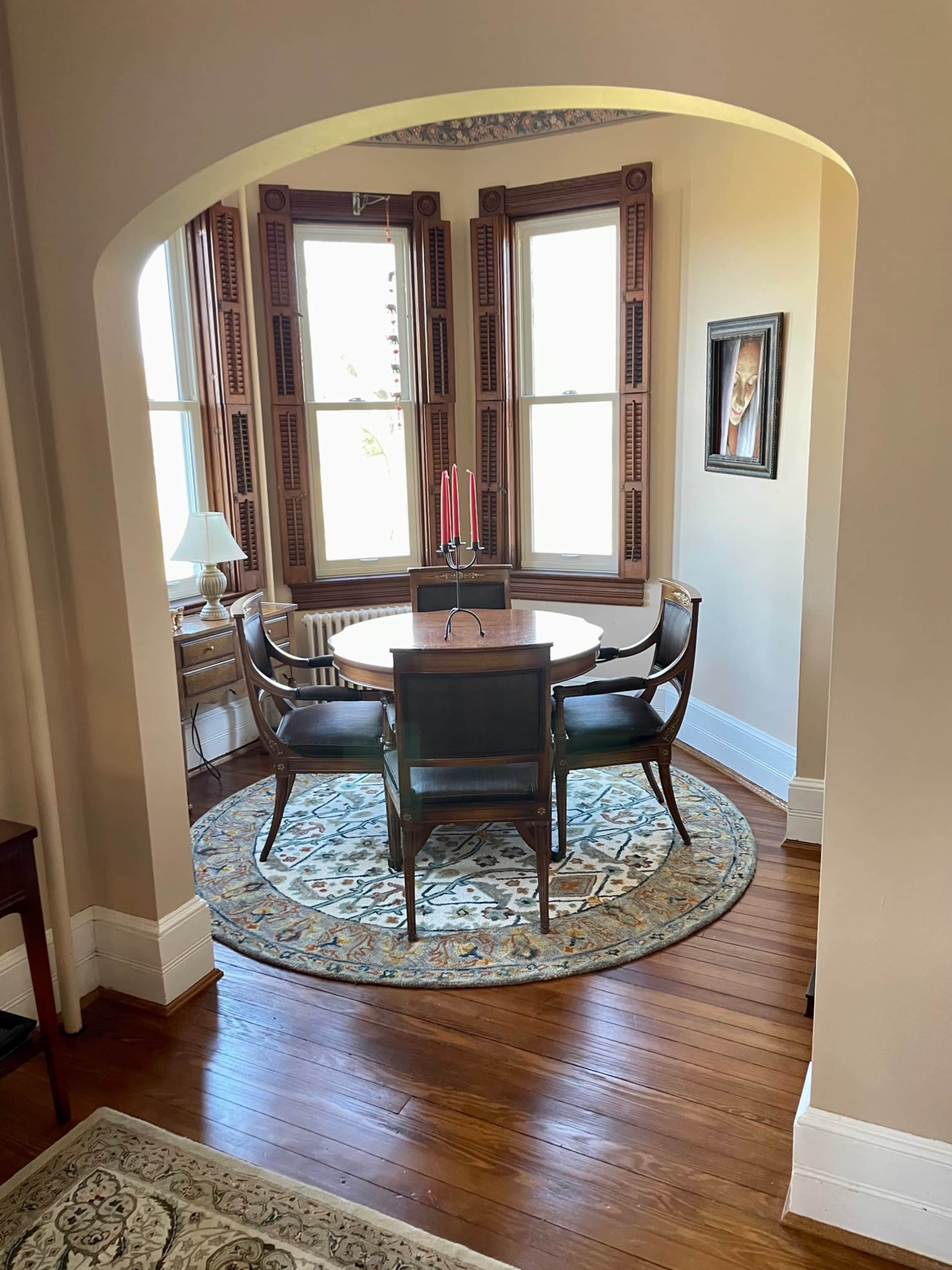 A dining area features a round table with chairs, situated on a patterned rug beneath a bay window with wooden shutters.