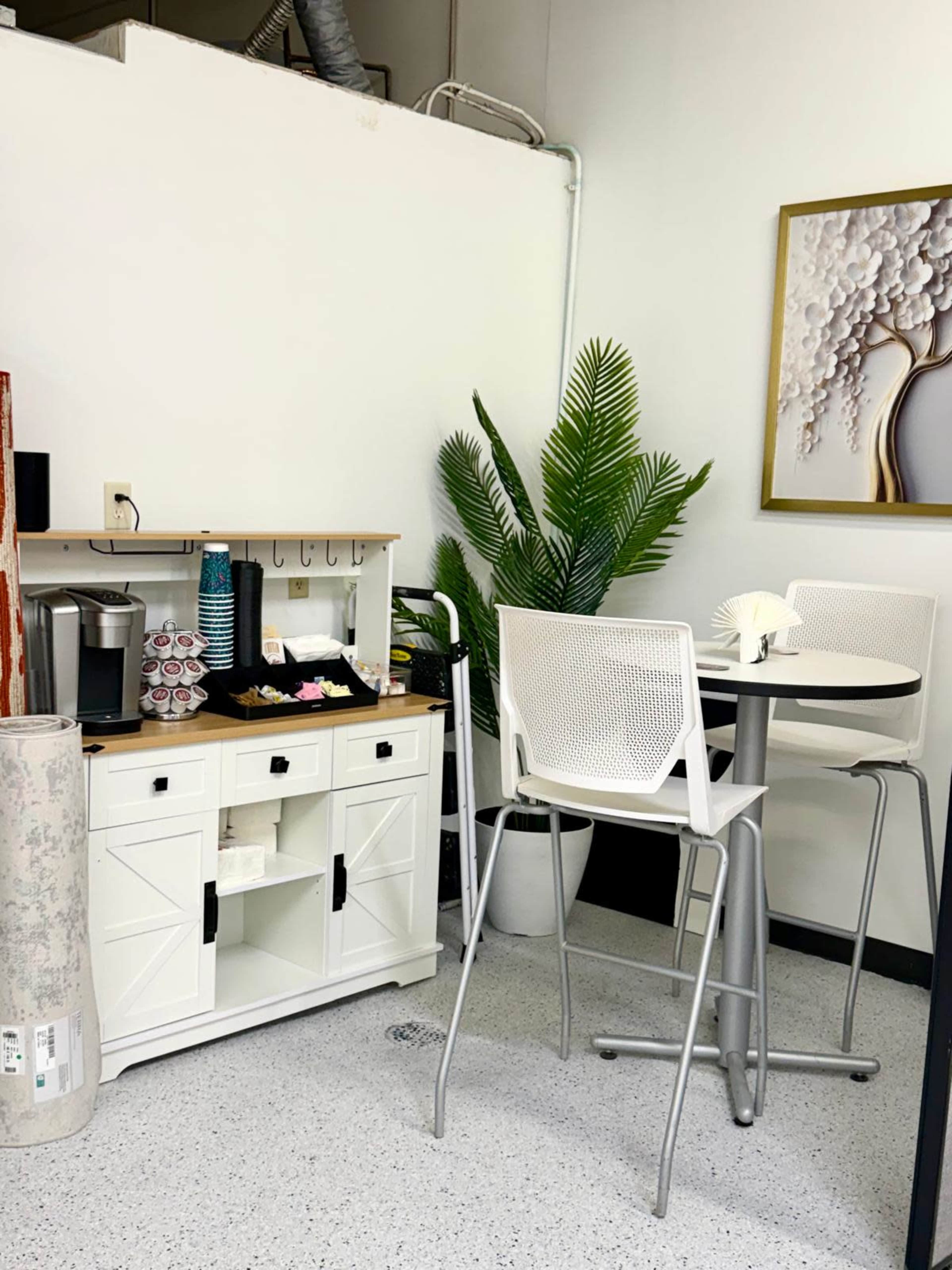 The image shows a small break area featuring a white wooden cabinet, a coffee machine, a round table with two chairs, and a potted plant.