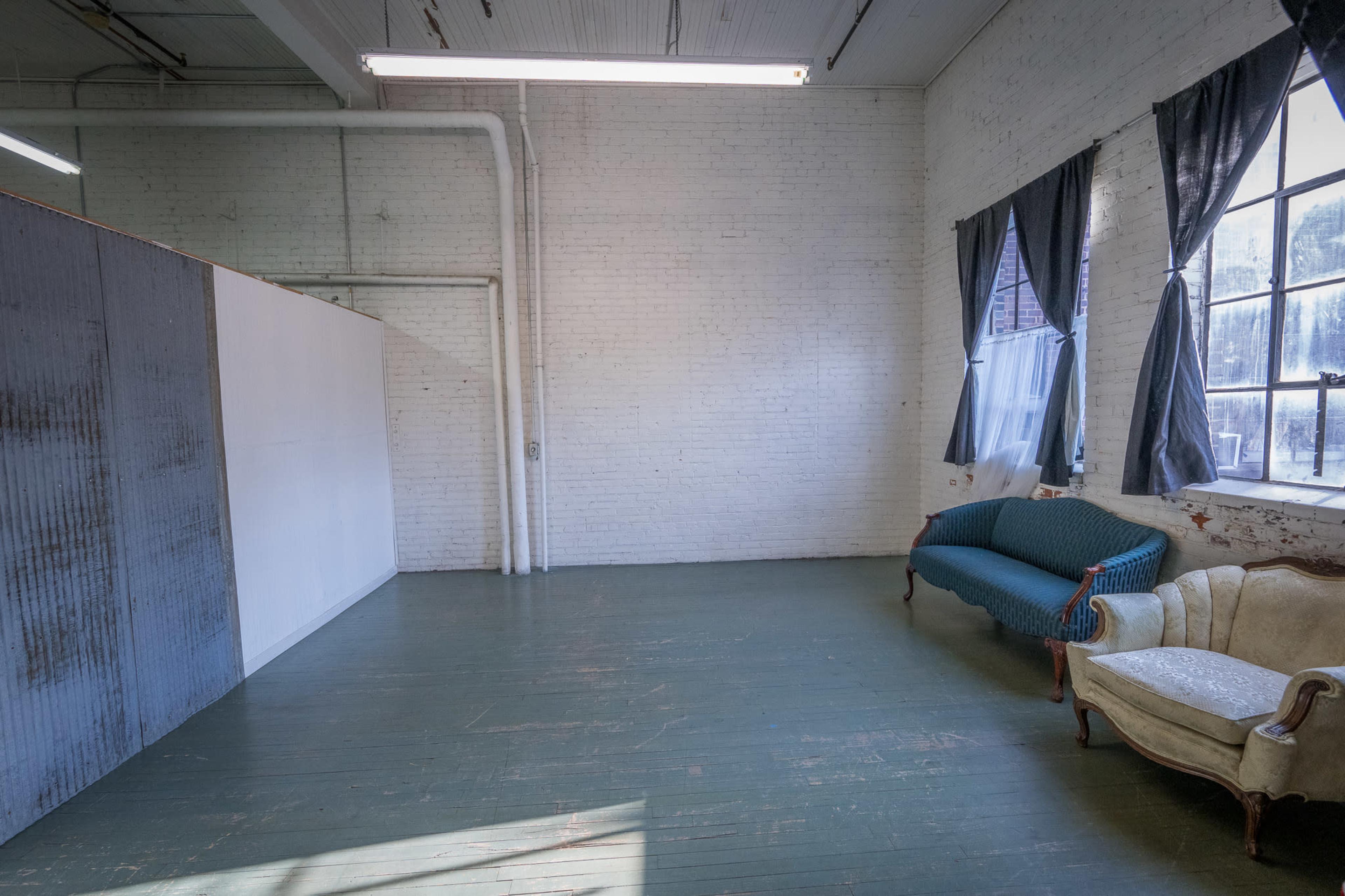 The image shows a spacious room with white brick walls, large windows covered by dark curtains, and two distinct sofas placed along one side.