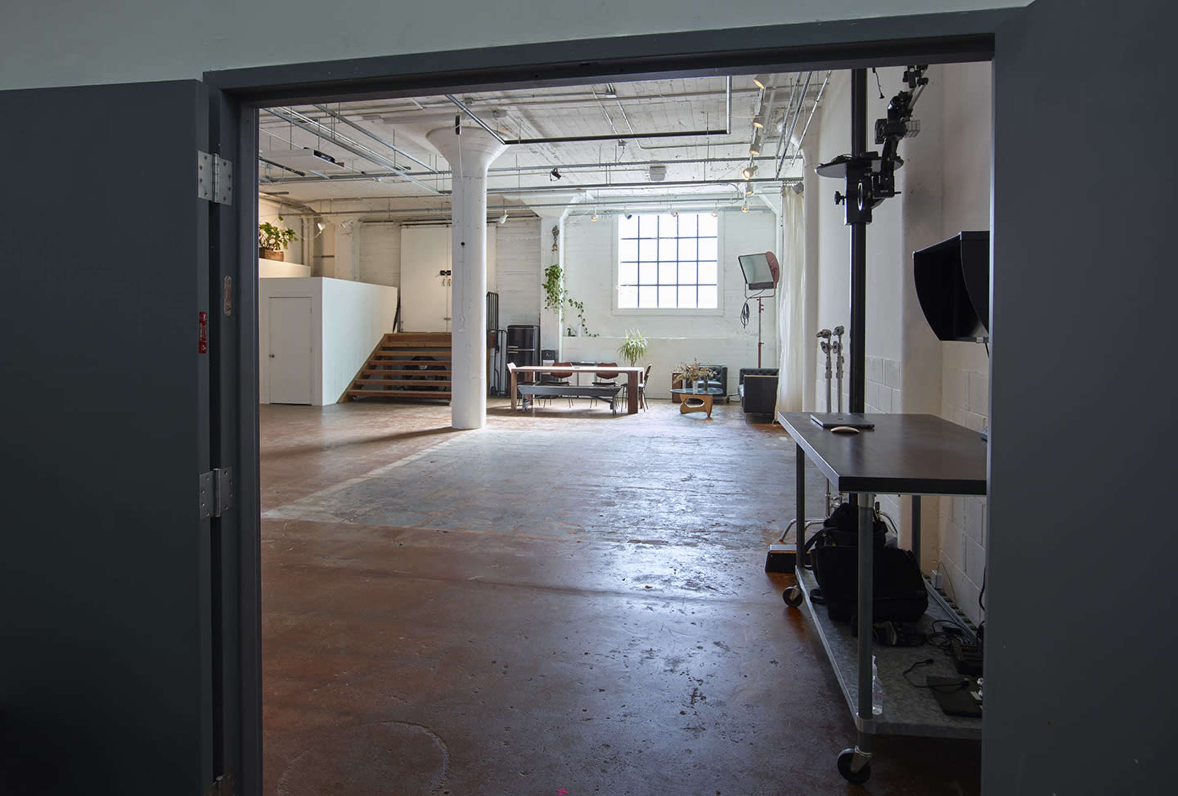A spacious, industrial-style room with concrete floors, large windows, and minimal furnishings, viewed from an open doorway.
