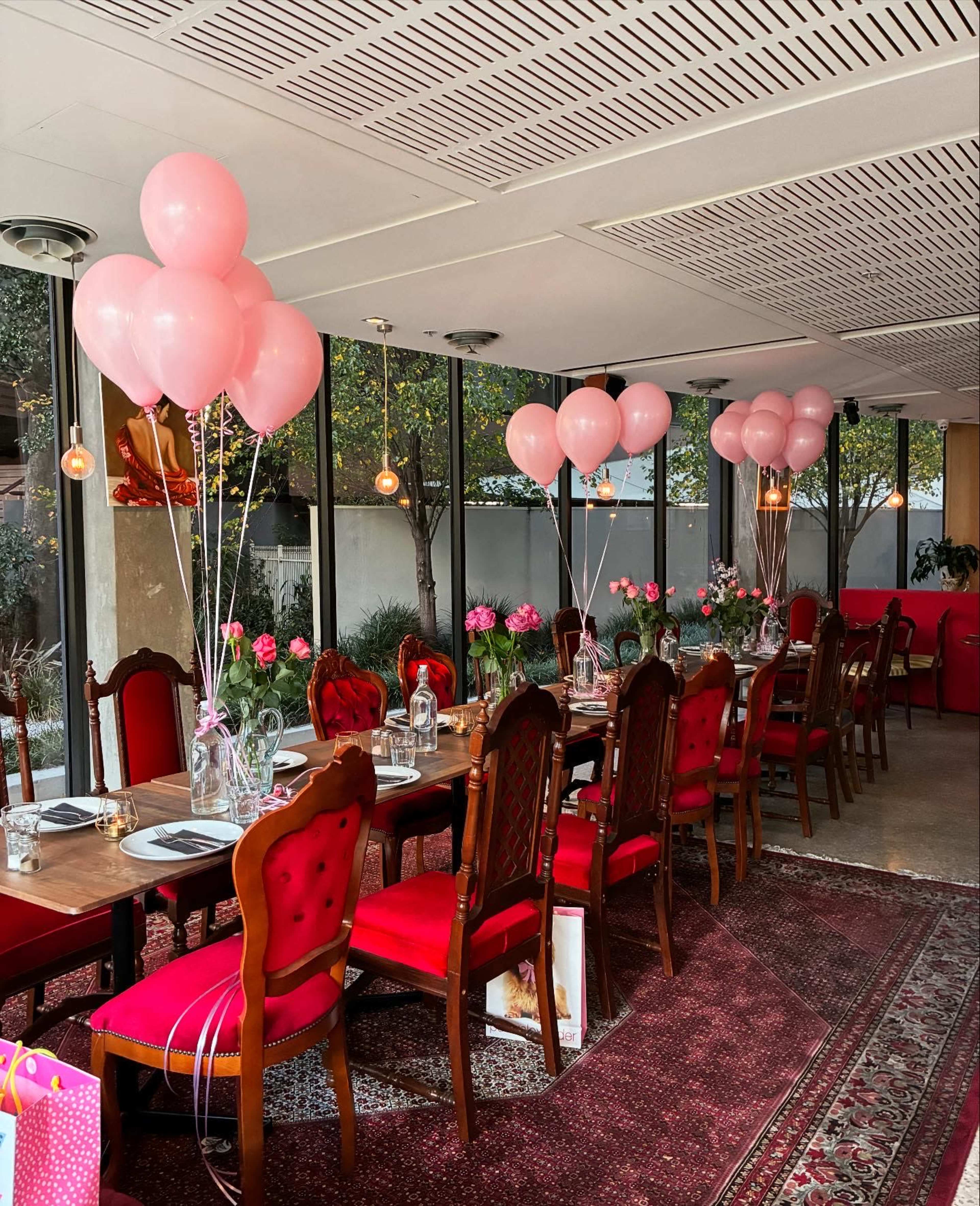 A long dining table is set in a brightly lit room, adorned with pink balloons and fresh flowers, surrounded by red chairs.