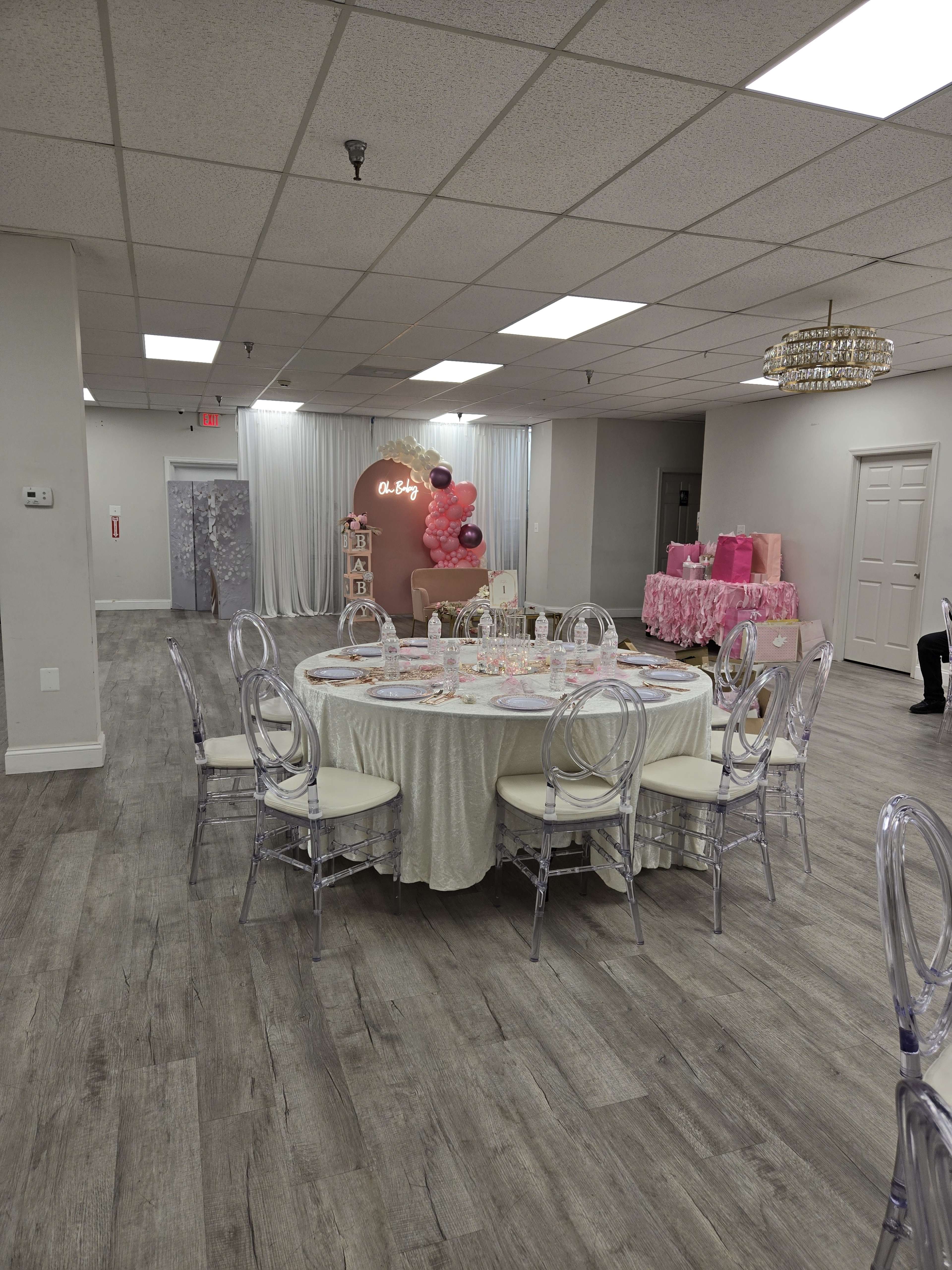 The image shows a banquet room set up with a circular table covered in a light-colored tablecloth surrounded by transparent chairs, with decorations and gifts in pink tones displayed in the background.