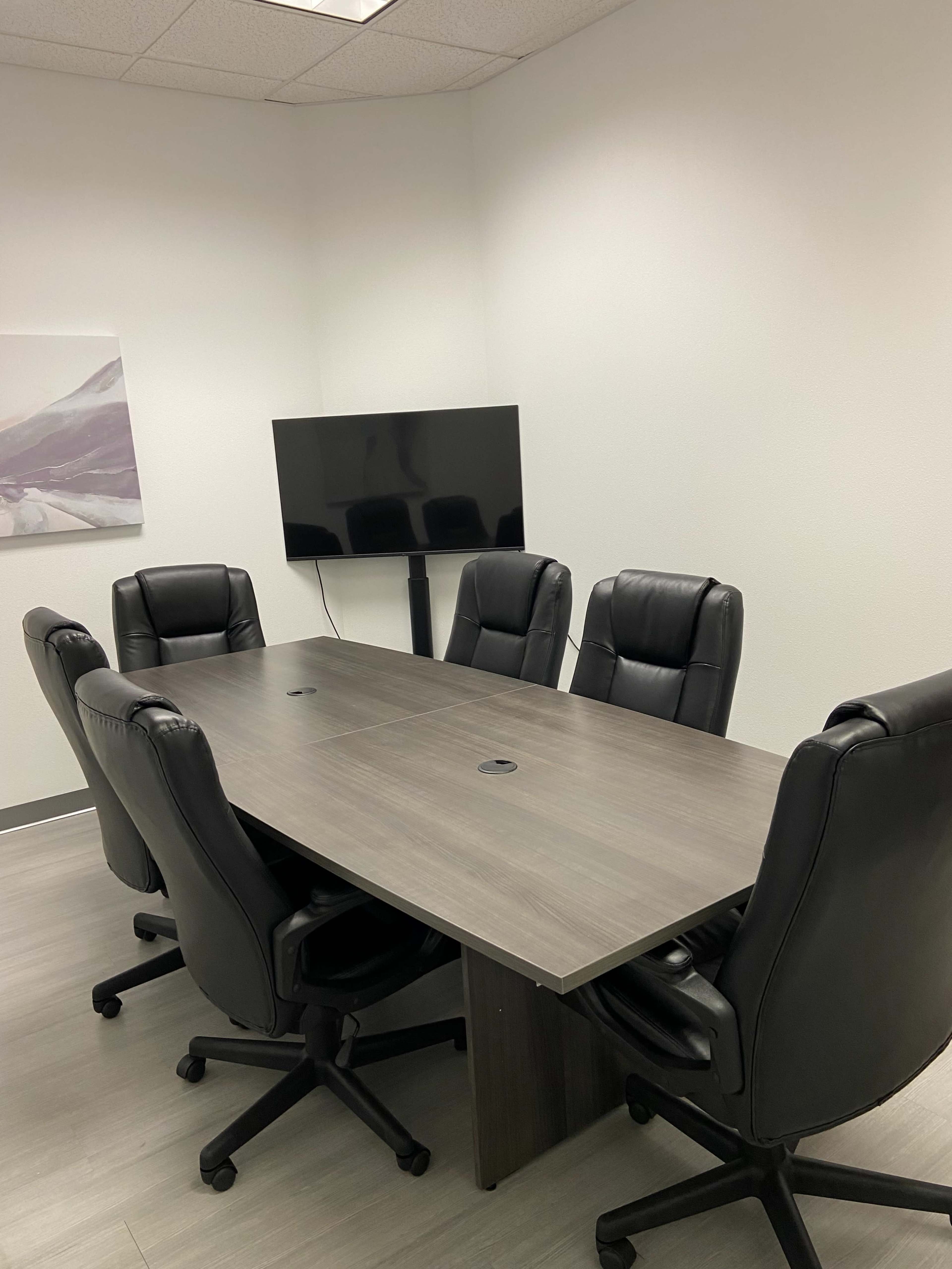 A conference room features a large rectangular table surrounded by six black ergonomic chairs and a wall-mounted television.